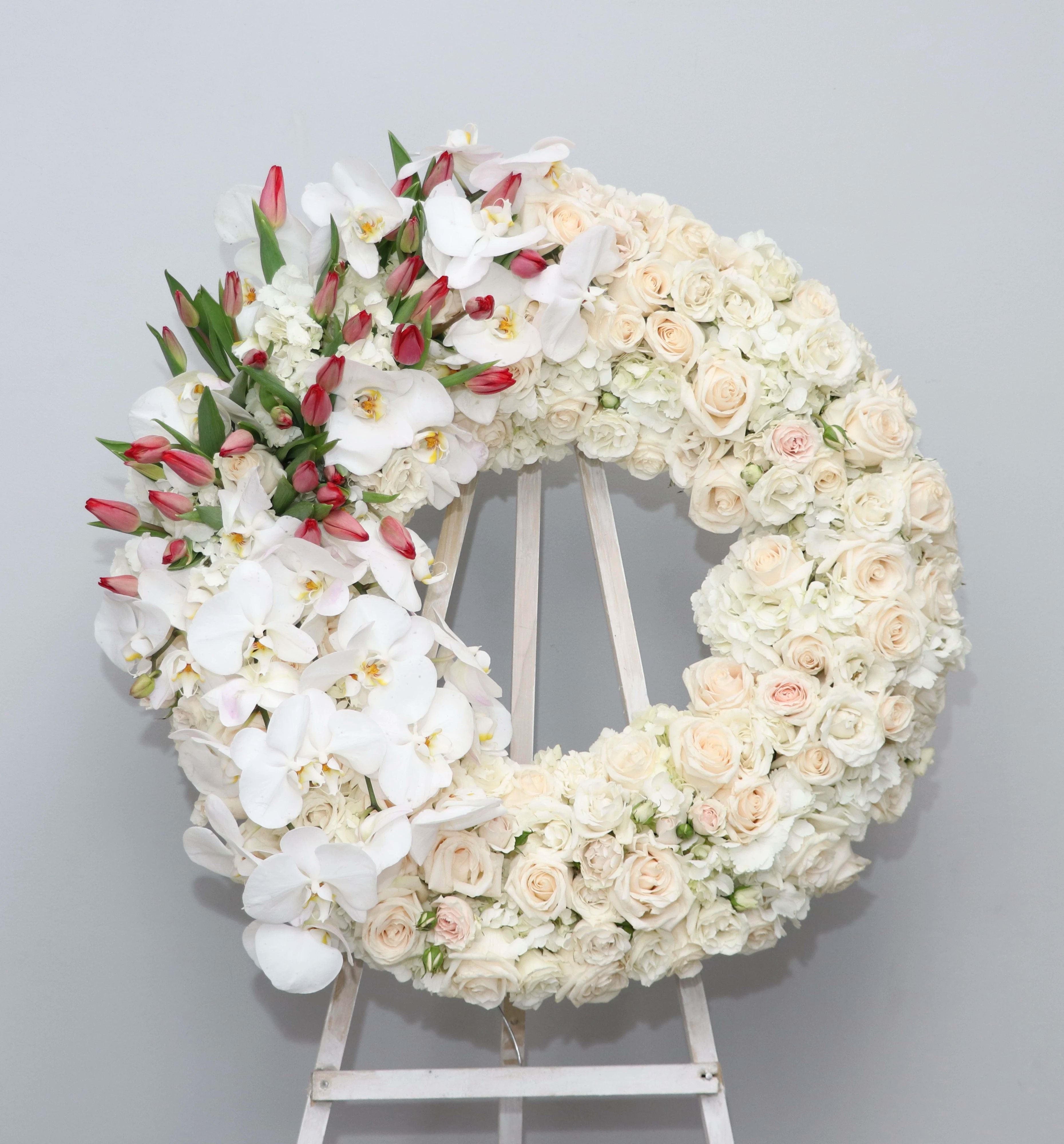 Etneral Flame Wreath - This elegant wreath is made with blush and white roses and accented with gorgeous phalaenopsis orchids and red tulips.     We include easel, printed banner and delivery (some fees may apply). An additional banner can be added for $15.    Standard size is 30'', deluxe is 36'', and premium is 42''.