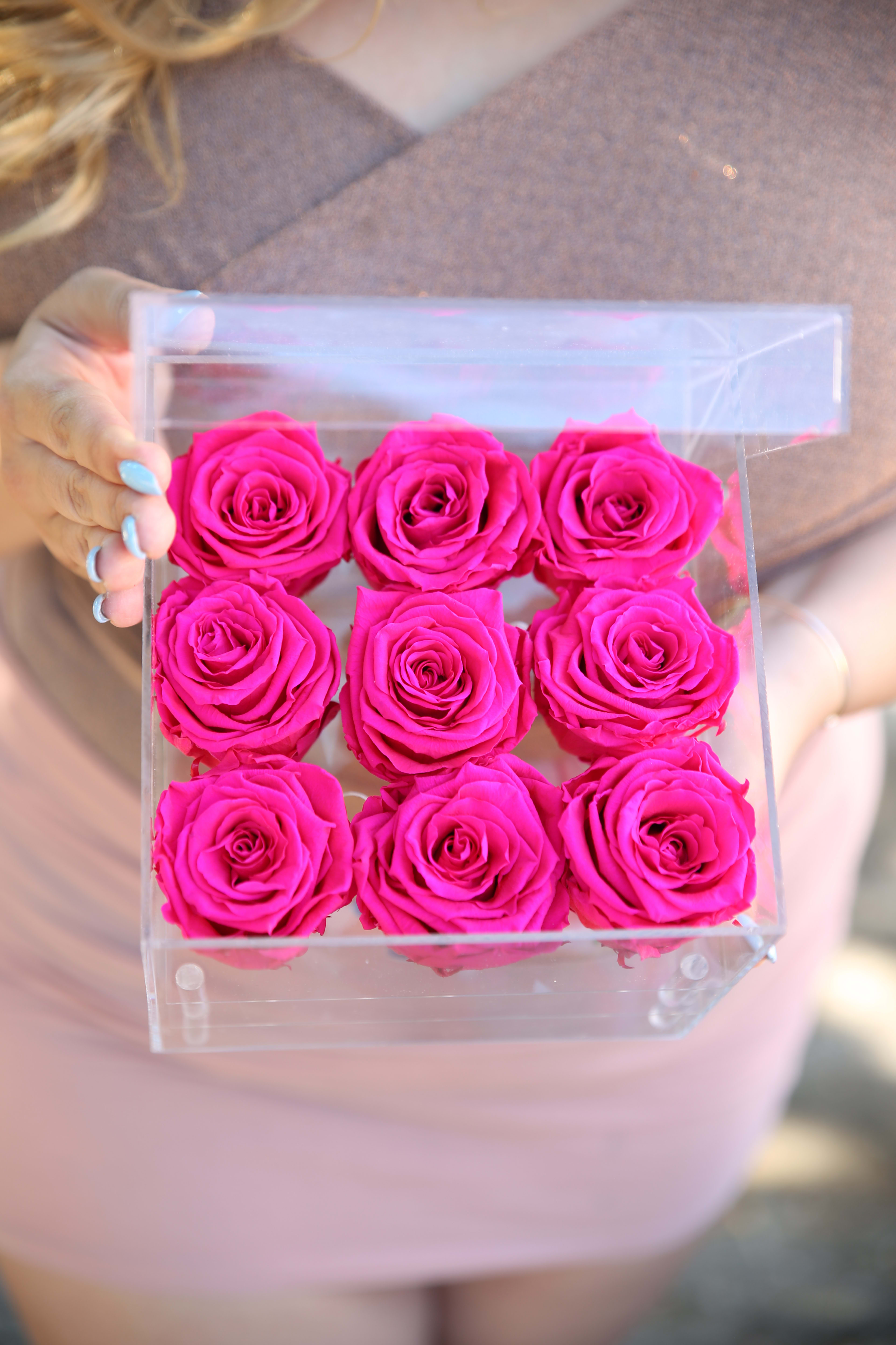 Small Clear Case - 9 One Year Roses in Hot Pink - Meet our everlasting roses! These are REAL roses that can last for up to a year! This case is composed of 9 Hot Pink Everlasting Roses. While supplies last, color subject to change based on availability.   About: Our everlasting roses, are imported directly from Ecuador where they are treated with a proprietary solution and color pigmentation process that transforms them into long lasting roses that maintain their freshness and biological structure. The roses are treated using a non-toxic and non-allergenic wax formula that is similiar to silica. The outcome is that our roses continue to look and feel like real roses - because they are real - with the difference being that they will maintain their integrity and color for up to a year.  Maintenance: The only maintenance they require is a light dusting from time to time. Do NOT water the roses. Do NOT remove the roses from their container. Do NOT place anything onto the roses as they can crush and damage them. Avoid direct exposure to sunlight. Keep the arrangement in a room temperature environment. Please note, the lid is meant to stay on the side or bottom of the box. Positioning dark colored roses next to light color roses may result in the darker rose color bleeding onto the light roses.