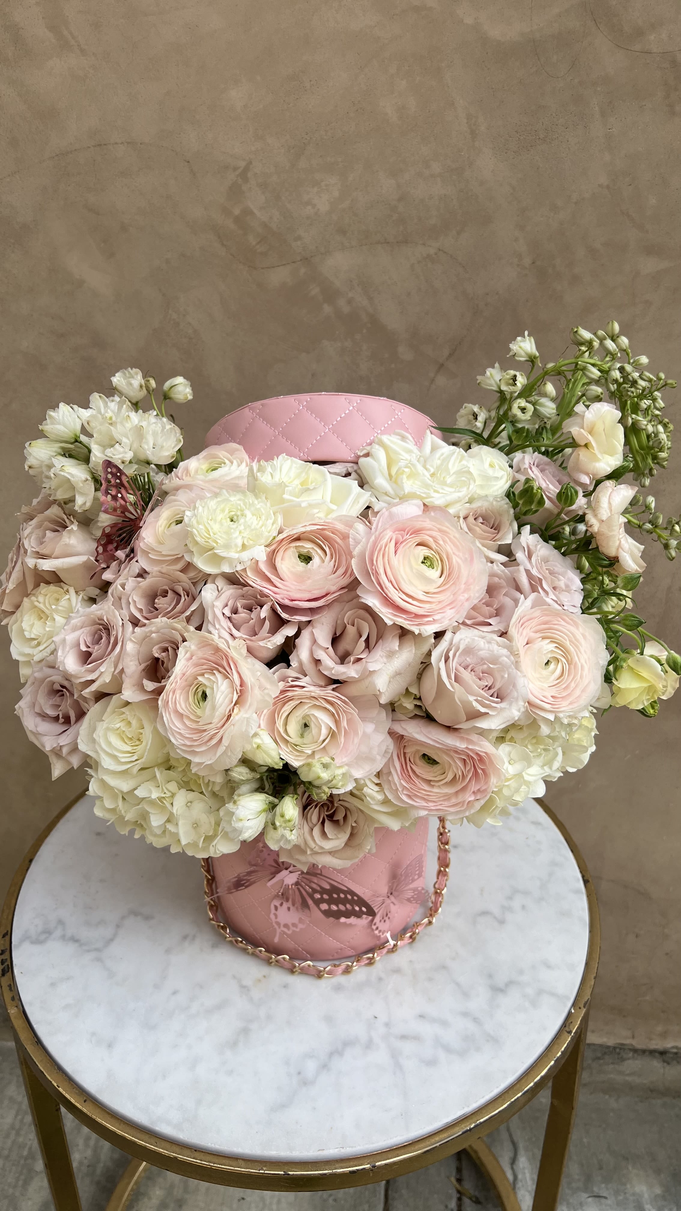 Blushing Butterfly Hat Box - The &quot;Blushing Butterfly Hat Box&quot; floral arrangement is a charming and delicate display of soft pinks and creamy whites, perfect for adding a touch of elegance to any occasion. This arrangement is nestled in a stylish pink quilted container adorned with a subtle butterfly accent. It's romantic and feminine aesthetic makes it an ideal choice for weddings, baby showers, or any event that calls for a touch of grace.  Please note that the flower varieties are subject to seasonal availability and may vary slightly to ensure the freshest and most beautiful blooms. Please note that the cushion hat boxes are available on a first-come, first-serve basis.
