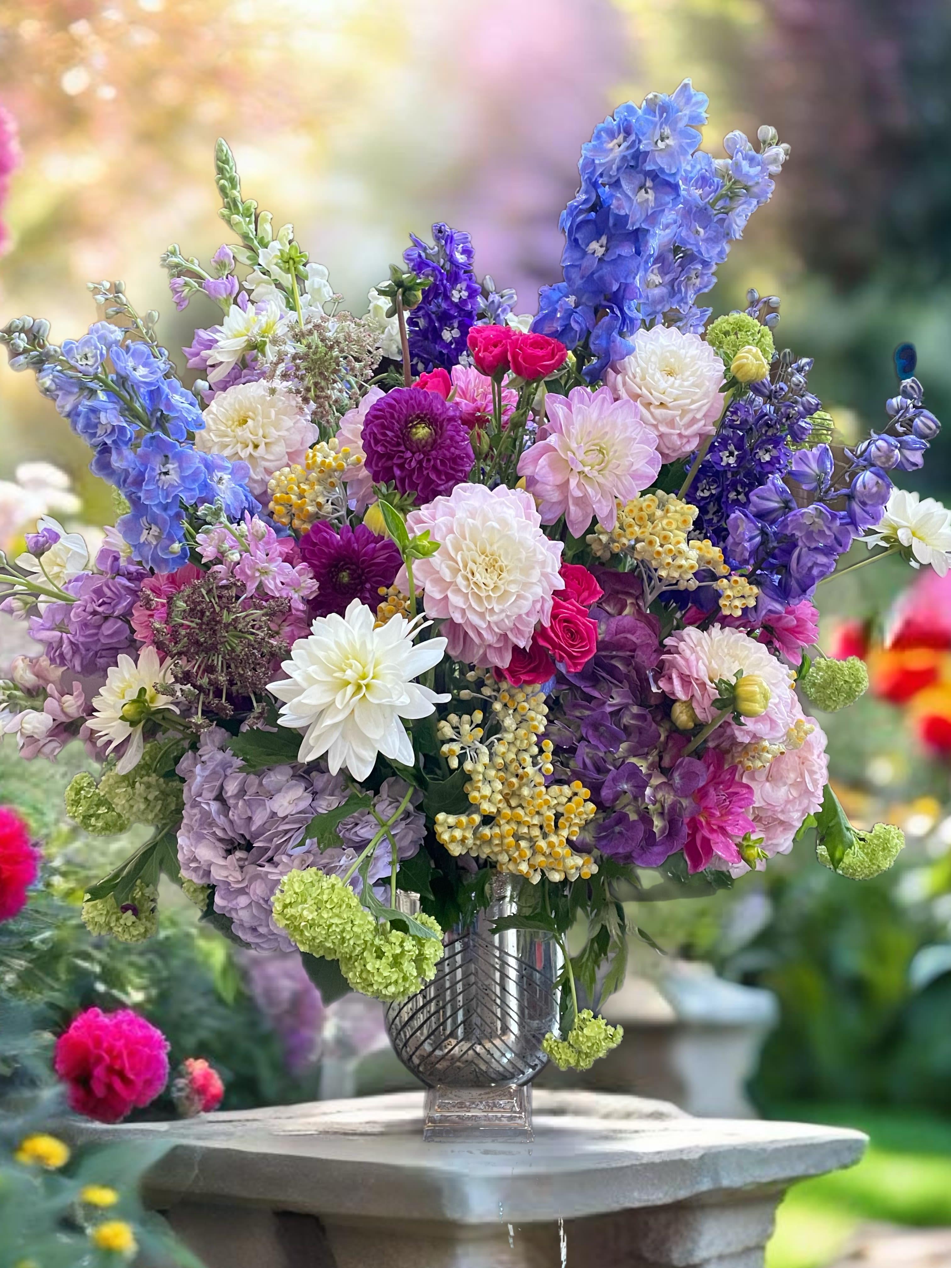 Fabulous celebration!  - breathtaking floral arrangement featuring vibrant dahlias in deep burgundy and soft blush, surrounded by elegant delphiniums in shades of blue and purple. Lavish roses, hydrangeas add texture and fragrance, while lush greenery like ferns and eucalyptus frames the blooms beautifully. This stunning display radiates sophistication and is perfect for any occasion.