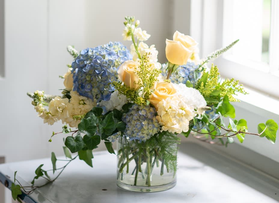Port Side - With colors like the sun reflecting on the water, these buttercream Roses and seasonal filler flowers pair charmingly with Blue Hydrangea and flowing greens. Florist reserves the right to make modifications to the design based on seasonality and accessibility of blooms and vases. Any modifications made will honor the palette, intent, and feeling of the pictured design.  Please kindly contact us directly with any specifications or concerns. 