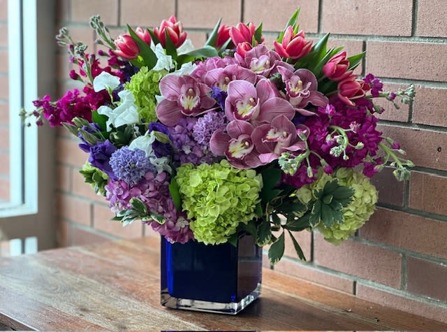 Radiant Blooming Garden Radiant Blooming Garden - Radiant Blooming Garden Bouquet by Venetian Flowers - The rich jewel tones bouquet is a vibrant and luxurious arrangement that captures the eye and the heart. At its core, lush green hydrangeas provide a fresh, verdant foundation, creating a stunning contrast against the bold colors that follow. Nestled among the hydrangeas are elegant pink cymbidium orchids, their exotic blossoms adding a touch of sophistication and grace. Bright tulips in a variety of jewel tones—deep purples, rich reds, and vibrant pinks—burst forth from the bouquet, infusing it with energy and life. Complementing these blooms, seasonal flowers in similar hues add texture and depth, creating a harmonious blend of colors that evoke the richness of a jewel box. All of this beauty is artfully arranged in a striking cobalt blue square vase, which not only enhances the vivid colors of the flowers but also adds a modern touch to the overall design. This bouquet is a captivating statement piece, perfect for any occasion that calls for a splash of color and elegance.
