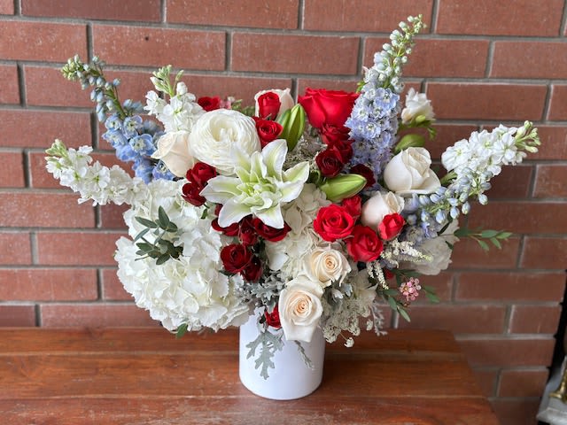 Stars And Blooms  - This striking red, white, and blue bouquet is a celebration of color and texture, artfully arranged in a sleek white ceramic cylinder vase. At the base, the fluffy white hydrangeas create a soft, voluminous foundation, providing a serene backdrop for the vibrant blooms that follow. Towering above, the elegant blue delphiniums add height and a pop of coolness, their bold spikes creating a beautiful contrast against the softer elements.  The bouquet features lush red roses and delicate spray roses, their deep crimson hues infusing the arrangement with warmth and romance. Interspersed among these are crisp white rose lilies, their large, fragrant blooms adding an air of sophistication, while the charming ranunculus adds playful softness with its layered petals.