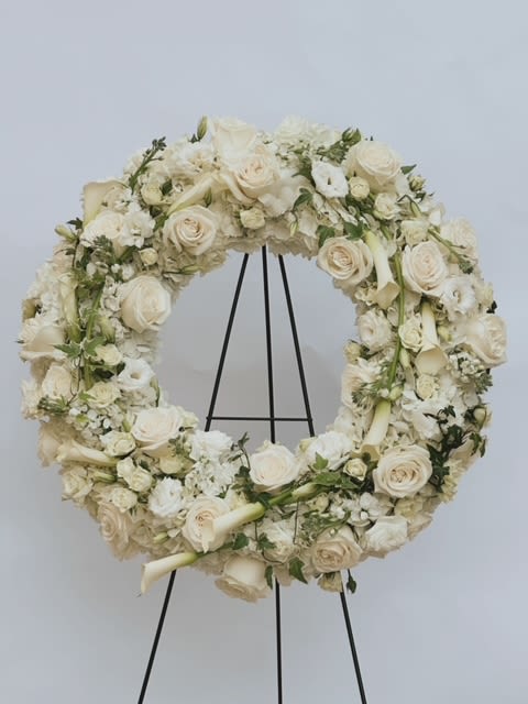 Heavenly Farewell Wreath By Venetian Flowers  - This stunning sympathy wreath on a stand is a poignant tribute, beautifully showcasing an array of white blooms that convey love, purity, and remembrance. The addition of white calla lilies elevates the design, enhancing its elegance and sophistication.  At the heart of the wreath, soft white hydrangeas create a plush, voluminous base, symbolizing heartfelt emotions and compassion. Surrounding these are classic white roses and delicate garden roses, their timeless beauty representing enduring love and cherished memories. The graceful white calla lilies, with their sleek and sculptural form, add a touch of refinement and symbolize beauty and resurrection, further enriching the meaning of the arrangement.  Interspersed throughout are white lisianthus and fragrant stock flowers, providing texture and a soft scent that enhances the overall experience. Other seasonal white blooms are thoughtfully included to create depth and a lush, full appearance. The circular shape of the wreath signifies eternity, making it a powerful symbol of everlasting love and remembrance. Shown in Standard 22&quot; Deluxe &amp; Premium will be designed on a larger 24&quot; base adding extra matching flowers - bigger grand look and feel 