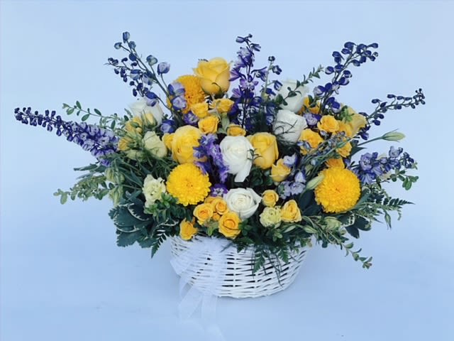 Sunlit Serenity Basket  - A charming white basket, filled to the brim with a vibrant arrangement of fresh-cut flowers. At the forefront, striking purple delphiniums rise tall, their deep hues creating a stunning contrast. Nestled among them are yellow and white roses, along with delicate spray roses that add layers of texture and beauty. Bright yellow ball mums bring a playful pop of color, while elegant lisianthus adds sophistication to the mix.  The arrangement is beautifully accented with lush greenery, providing depth and a refreshing touch to the overall design. This delightful creation by Venetian Flowers exudes warmth, making it a perfect gift for a variety of occasions. 