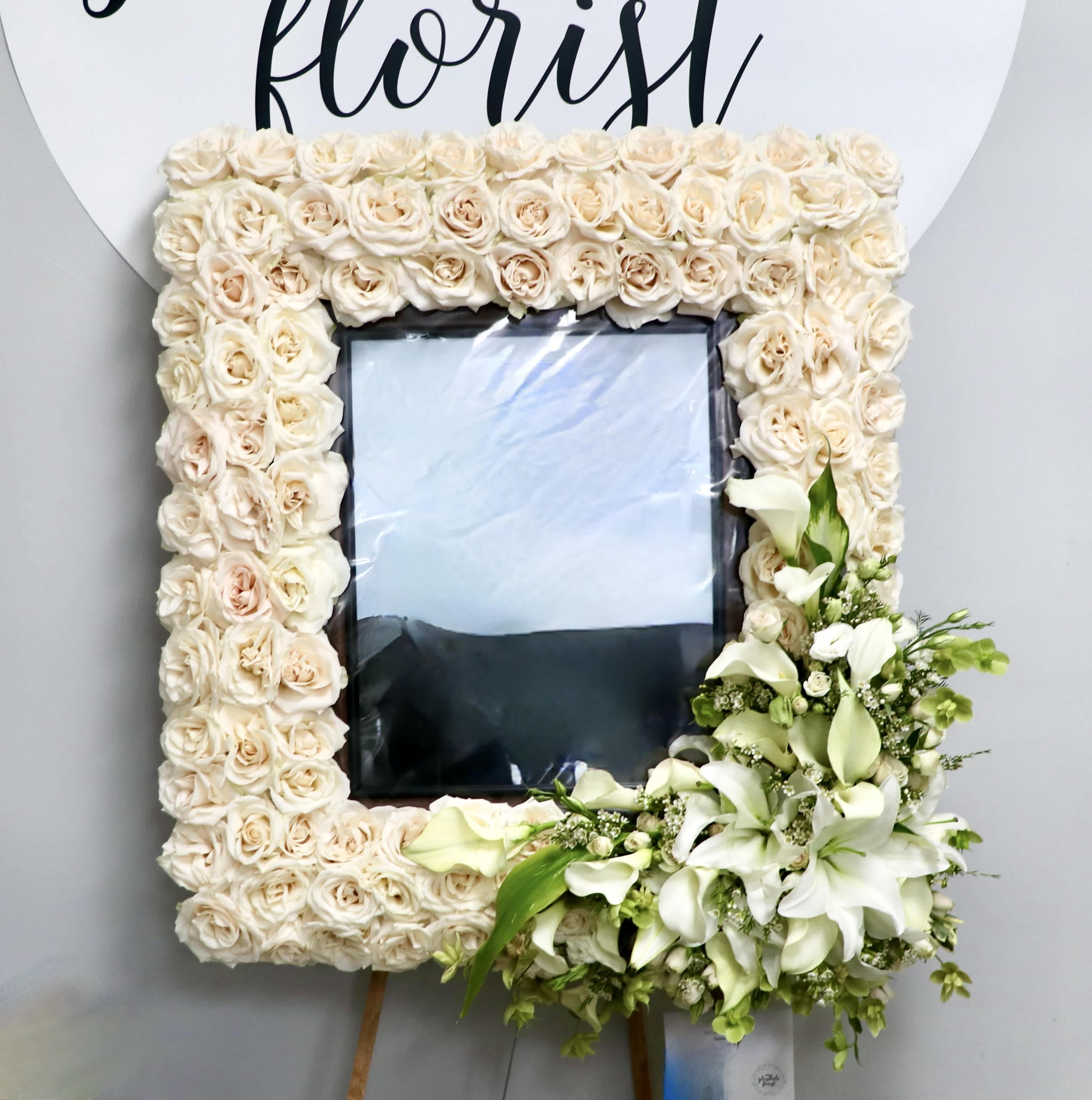 Rose and Lily Frame Rose and Lily Frame - This white sympathy frame is made with all white roses and a lily and greenery corner accent. This frame is a lovely choice to honor your loved one. Standard is about 32" wide, deluxe 36", premium 42" 11" x 14" frame is not included, please coordinate with us for drop off We include easel, printed banner, and delivery (some fees may apply). An additional banner can be added for $15