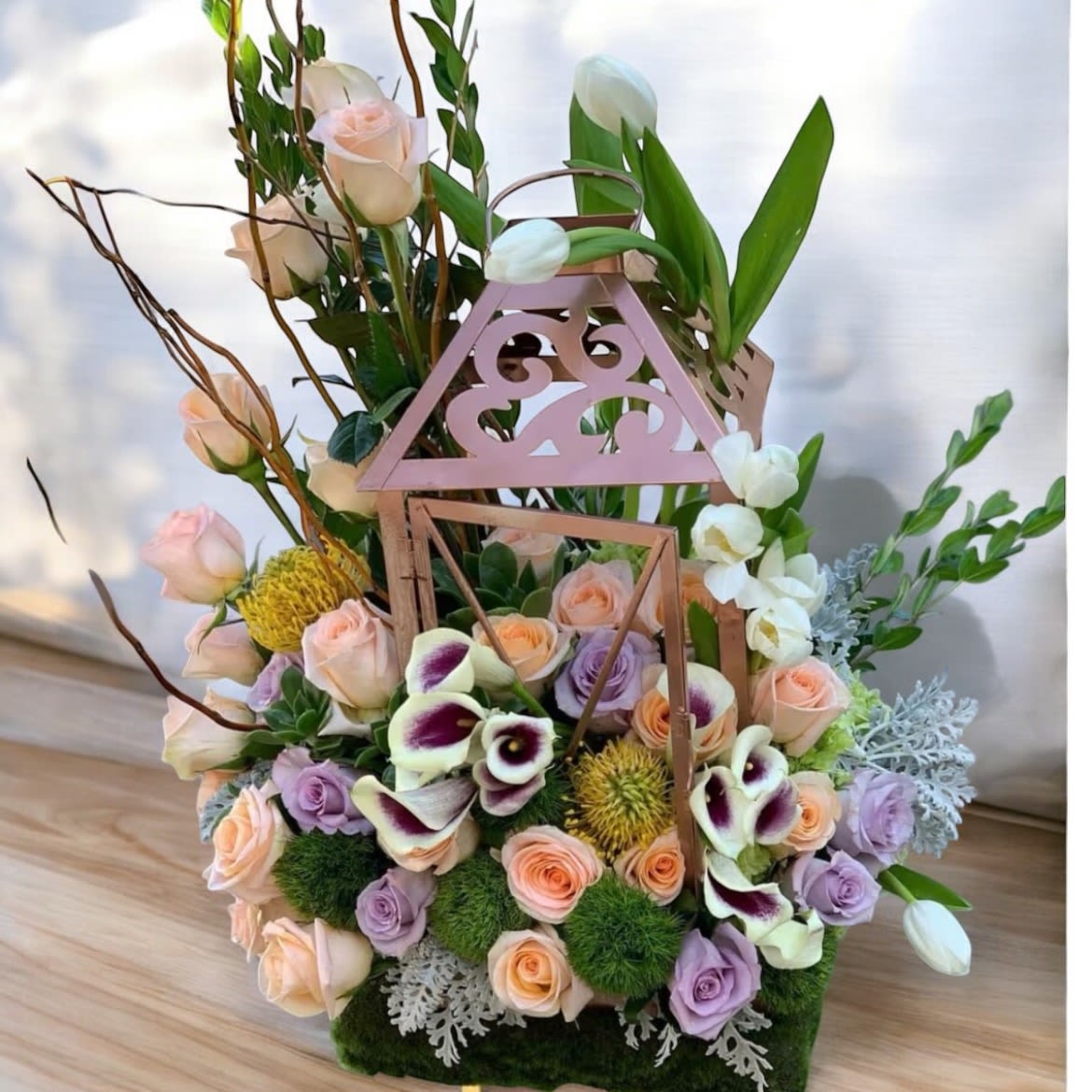 Lantern Garden Vibes Lantern Garden Vibes - Unique arrangements in one of a kind lantern! This arrangement includes roses, Calla lily, tulips, succulent, green moss, pincushion protea, cult willow, dusty miller. Lanterns available in different colors upon request.