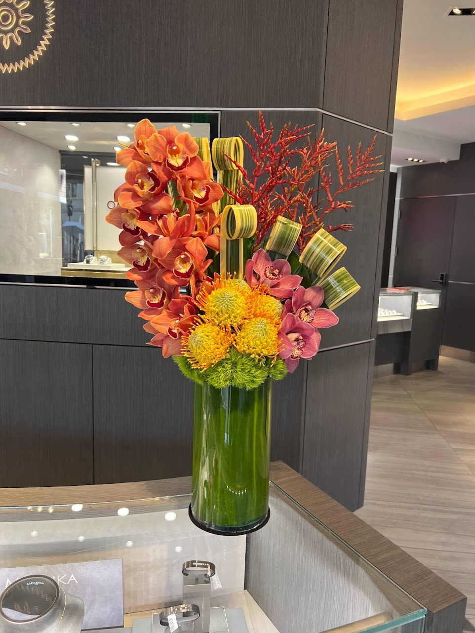 Tropical Paradise -  A vibrant floral arrangement featuring orange orchids, pink orchids, yellow pincushions, and green foliage, all elegantly arranged in a vase.   