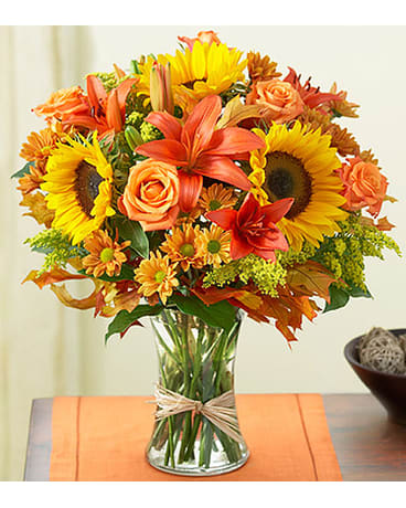 Fields Of Europe for Fall Fields Of Europe for Fall - Arrangement of orange roses and lilies vibrant sunflowers poms and more hand-gathered in a classic glass vase and finished off with charming raffia ribbon. All flowers and colors are subject to change due to availability. FCF-0067
