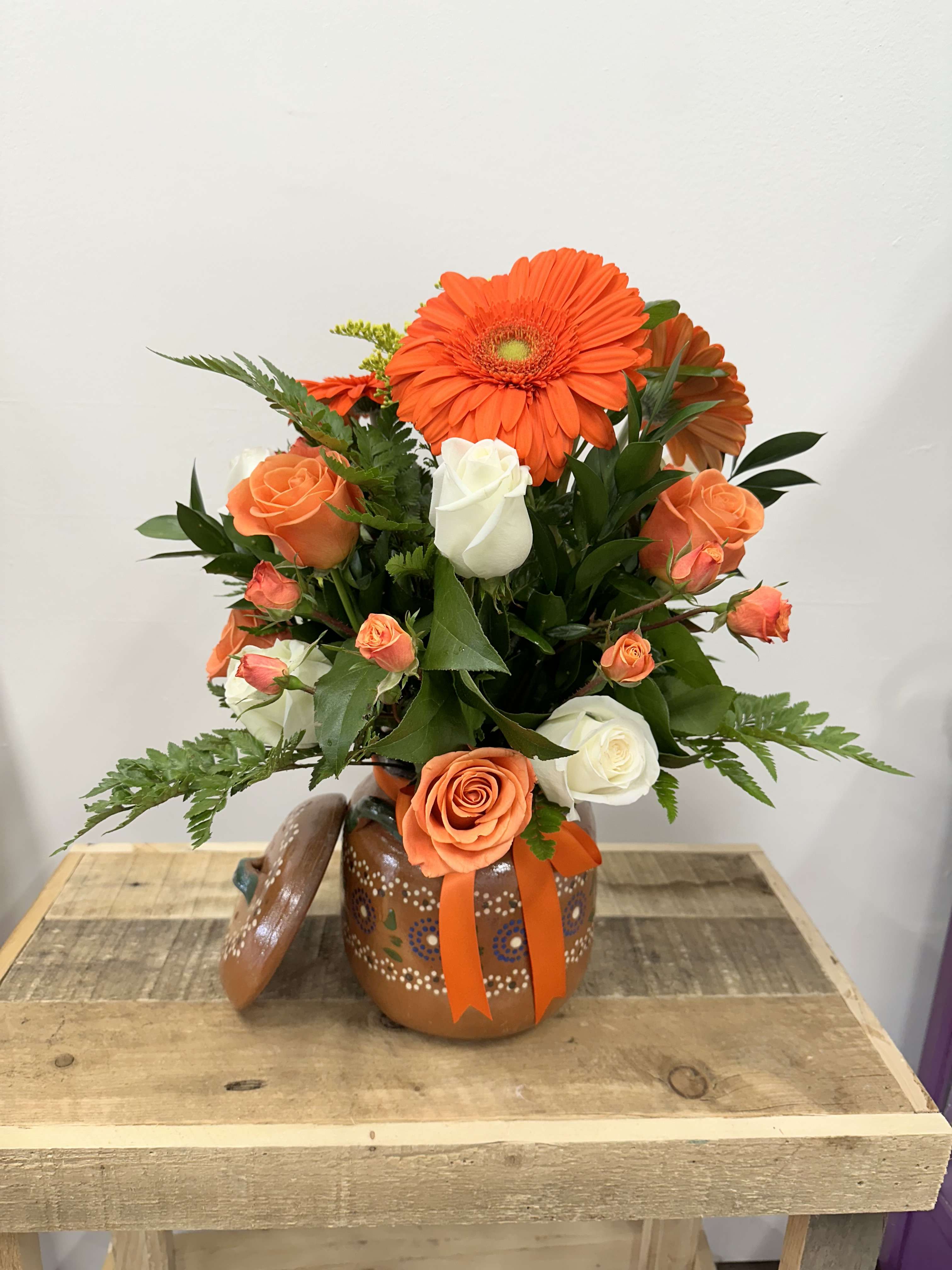 Sunrise Serenade Olla - A striking clay or arrangement featuring a vivid mix of white and orange roses, bright orange daisies and lush orange spray roses, all complemented by a touch of fresh greenery This bold and lively display combined vibrant colors, and Rick’s textures bring a warm and inviting setting.