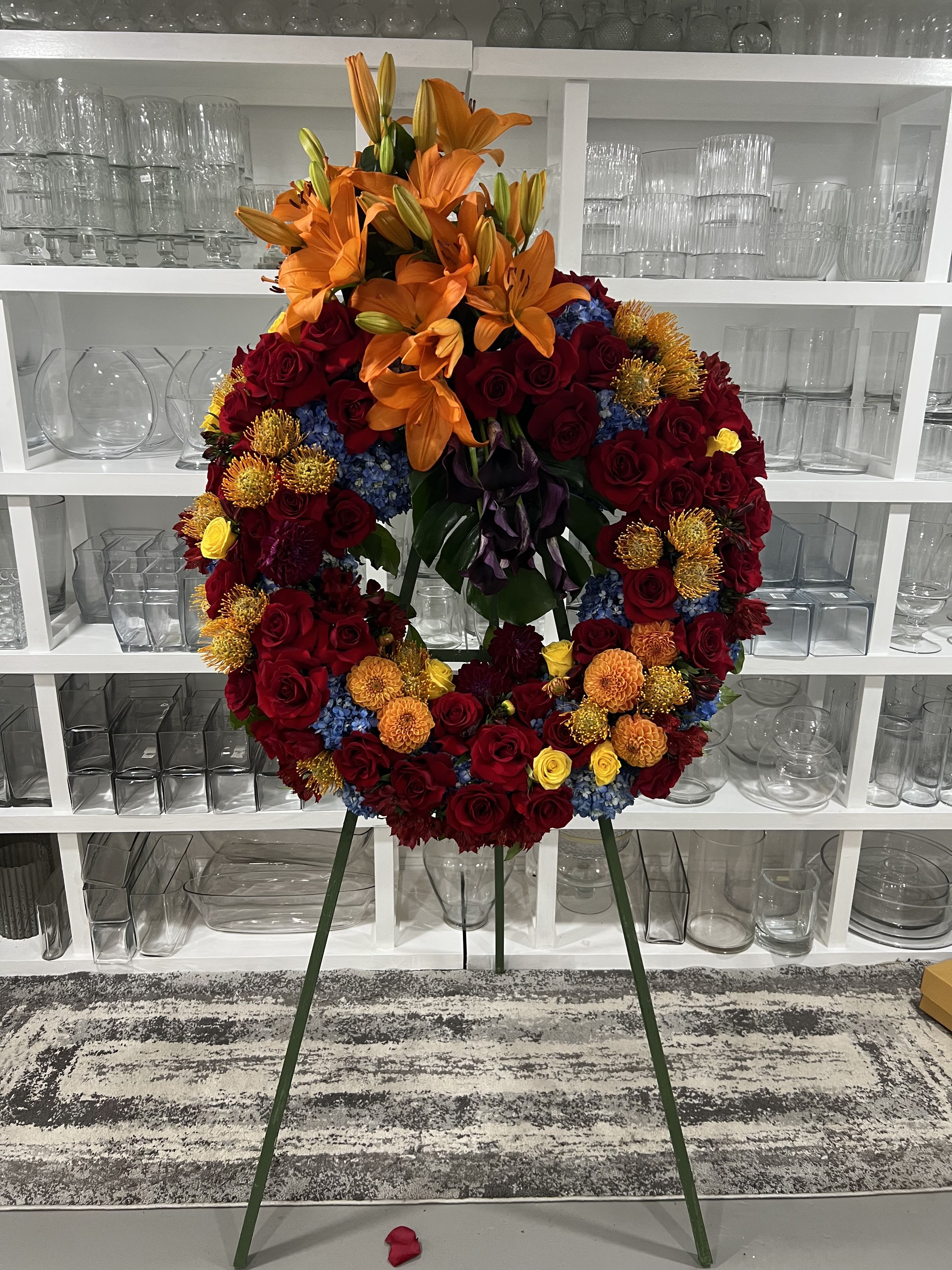 Eternal Garden Wreath - The &quot;Eternal Garden Wreath&quot; is a dignified and elegant floral tribute, carefully crafted to express deep respect and remembrance. This standing wreath arrangement features a harmonious blend of vibrant orange lilies and deep red roses, symbolizing the enduring beauty of life and love. Complementing these are purple calla lilies, which add a touch of serenity and grace. Bright yellow roses and blue hydrangeas introduce a sense of peace and calmness, reflecting cherished memories. Set on a discreet and sturdy metal stand, this wreath is a profound and heartfelt expression of sympathy, suitable for honoring a loved one at a funeral or memorial service.