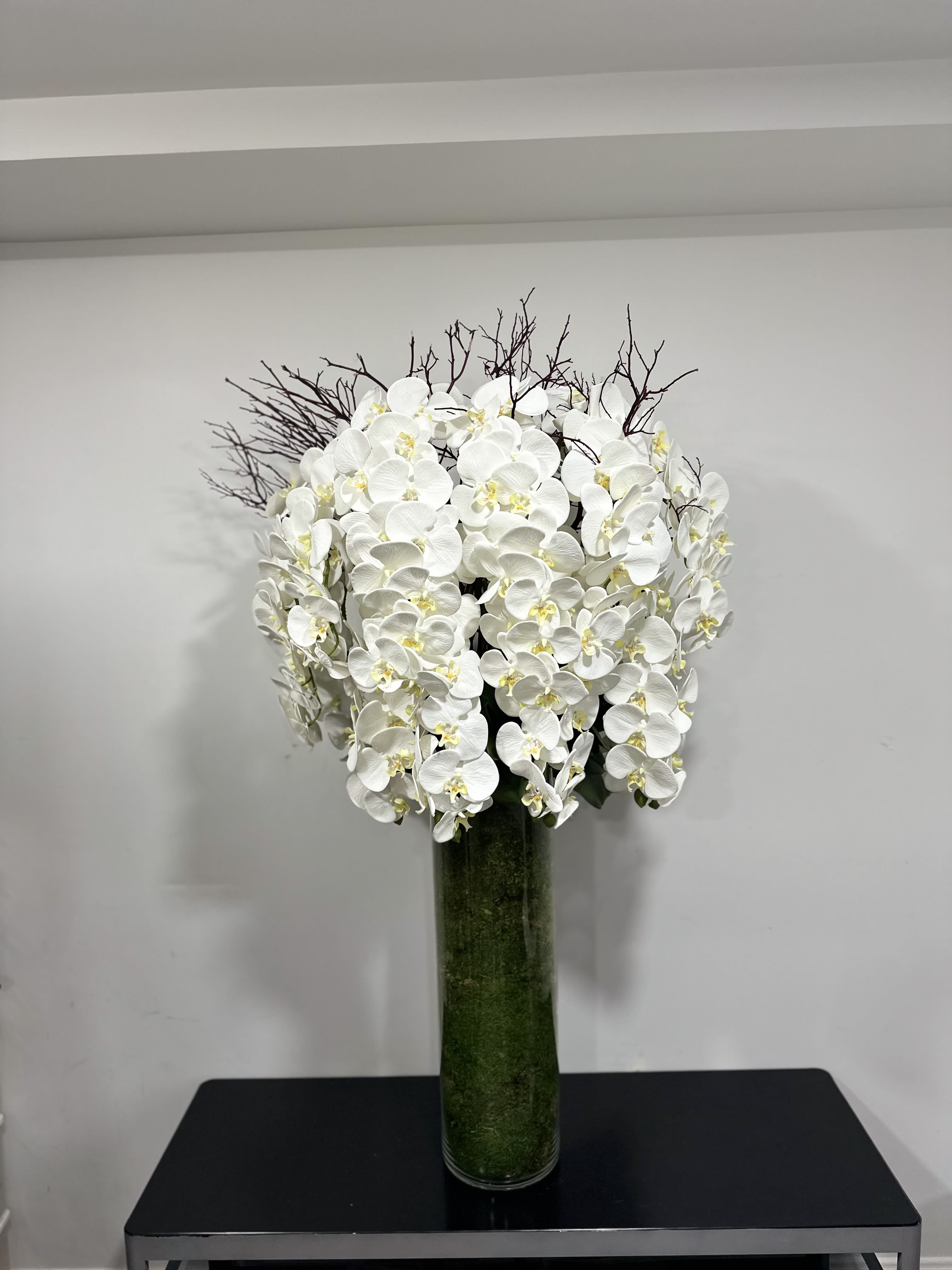 Celestial Grace - This exquisite floral arrangement, named &quot;Celestial Grace,&quot; features a captivating ensemble of pristine white silk orchids, each petal meticulously crafted to radiate a serene elegance. Nestled amidst a dramatic array of slender, dark twigs, the orchids stand out with an almost luminous quality, evoking a serene, celestial atmosphere. The arrangement is thoughtfully placed in a sleek, transparent glass vase, filled with lush green moss that adds a touch of earthy vitality while grounding the lofty floral display. &quot;Celestial Grace&quot; is a testament to sophisticated design, perfectly suited for enhancing the decor of any refined setting, offering a visual retreat that beckons a moment of quiet admiration and contemplative beauty.