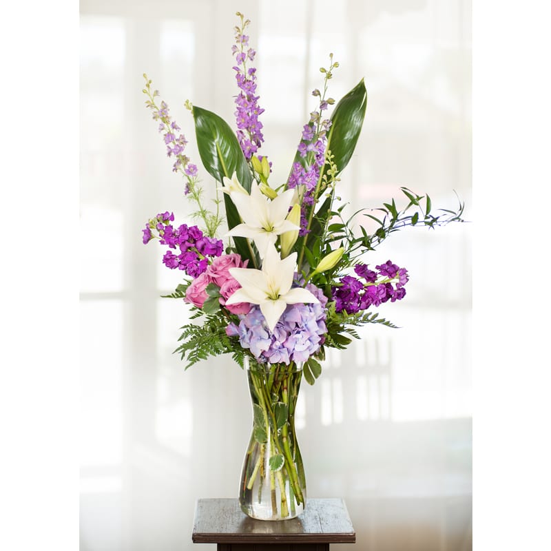 Purple Wishes***** Purple Wishes***** - Item Code: 109-VF The Purple Wishes bouquet is the perfect gift for any occasion. The unique glass vase is filled with stock, larkspur, hydrangea, and roses in different shades of purple. White Asiatic lilies are set in the center of this eye-catching design. approx. 32"h x 10"w