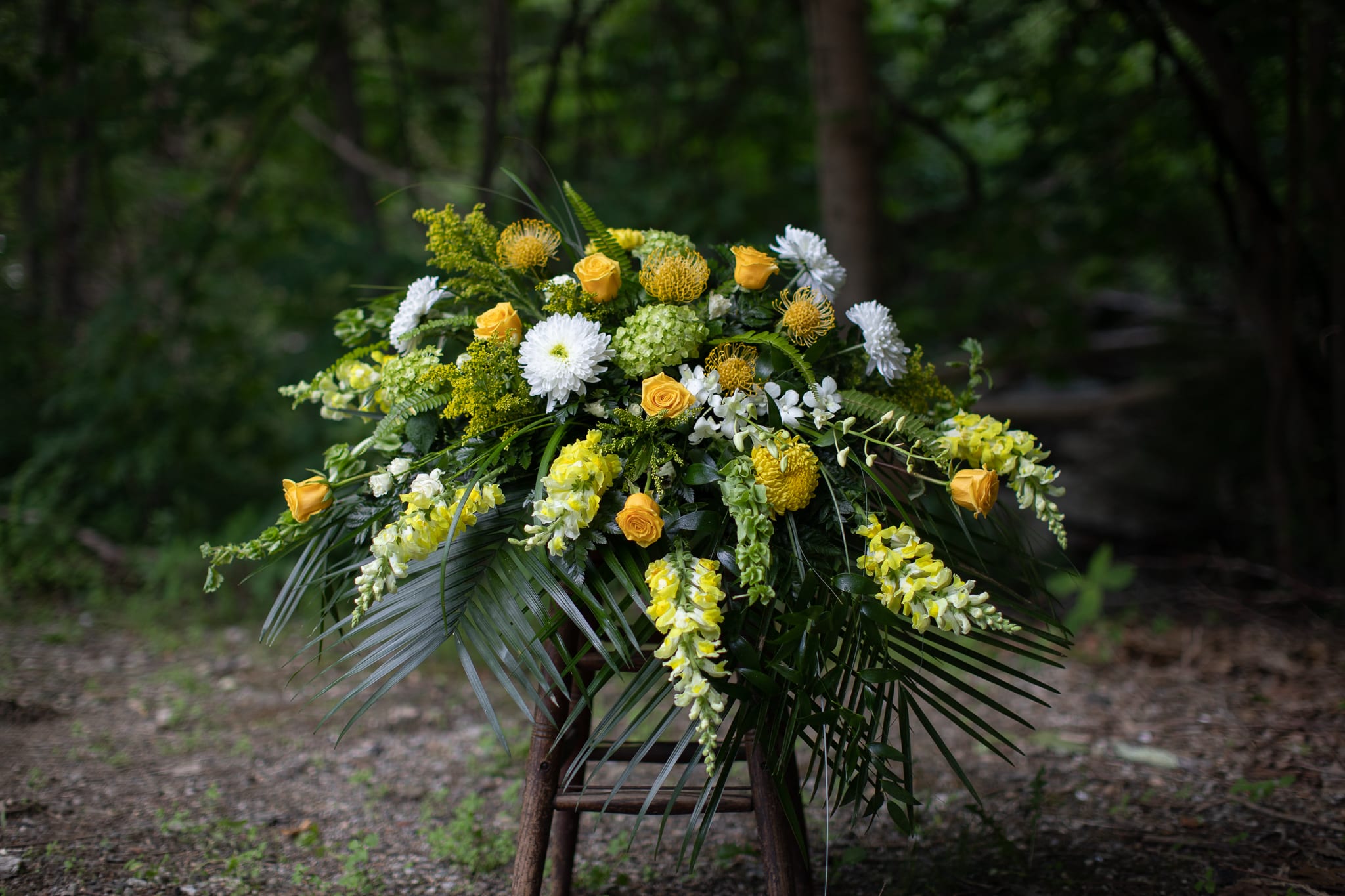 Golden Memories Casket Cover - A sunshine sentiment created with golden yellow and ivory toned seasonal flowers. Blooms are selected by seasonality. Focal flowers may include Asiatic lily, sunflowers or roses with bountiful hydrangeas, snapdragons and other wildflower blooms. A warm and loving send off for those we hold near and dear in our hearts. Please call with special requests. 