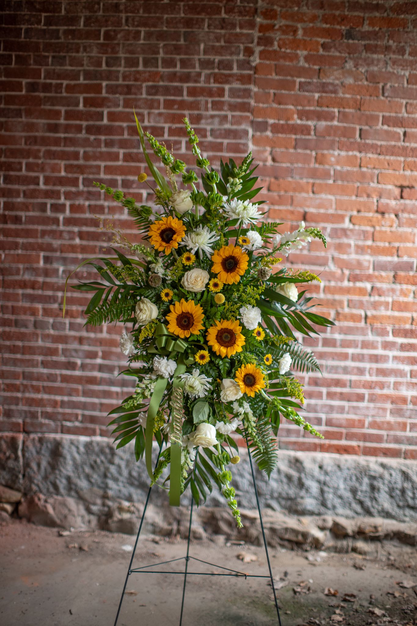 Golden Memories Hanging Spray - A warm send off to honor a life well lived an loved. This floral design has a free floating effect. It is placed on an easel to showcase each  bloom. Flowers are arranged in a geometric pattern that mimics a fire work. Floral selections will be in golden, yellow, cream, white and green tones. Please call with special floral requests or give us twenty four hours notice to be sure we can source the flowers you're looking for. We do not always have sunflowers during the winter months/season. 