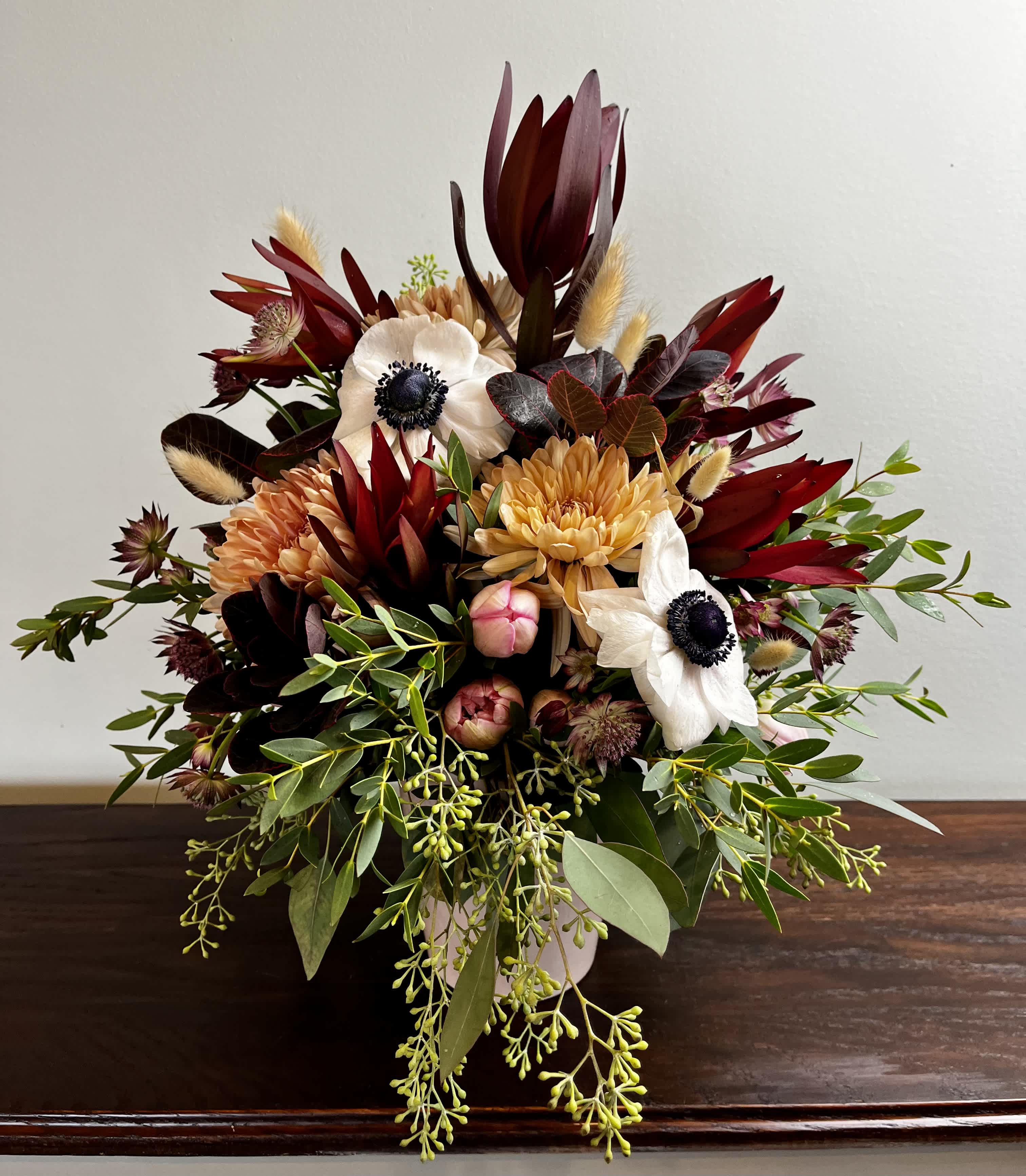 Lavishly Lush - A rich display of colors and textures.  Including Anemones, Ruby Leucadendrons, Peach Cremons, Pink Tulips, Astrantia, Bunny Tails, Smoke Bush, and mixed greens in a pink ceramic vase.   