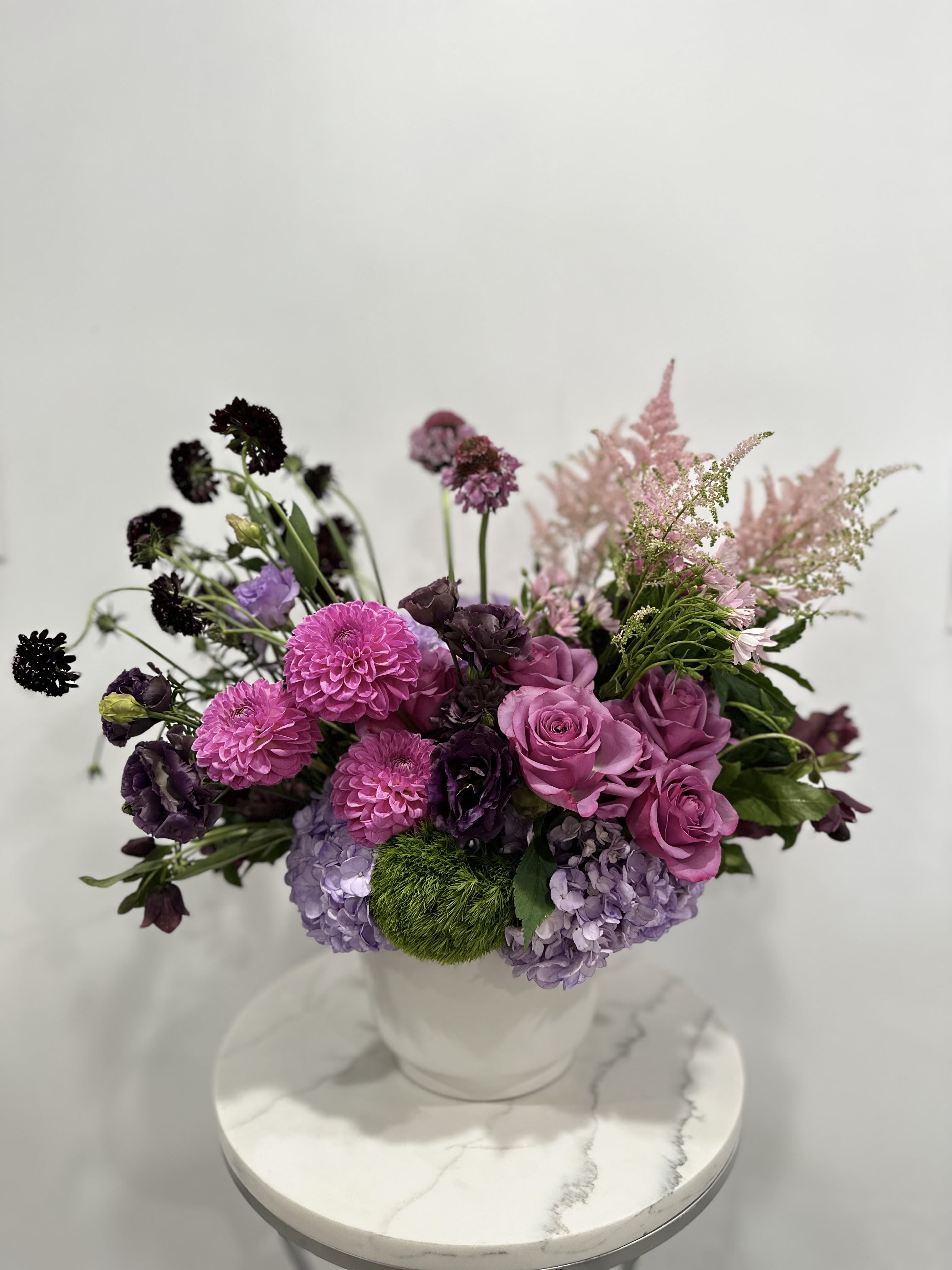 Lavender Bliss - Elegant floral arrangement with vibrant pink dahlias, lavender roses, and deep purple accents, complemented by soft greenery in a chic white vase.
