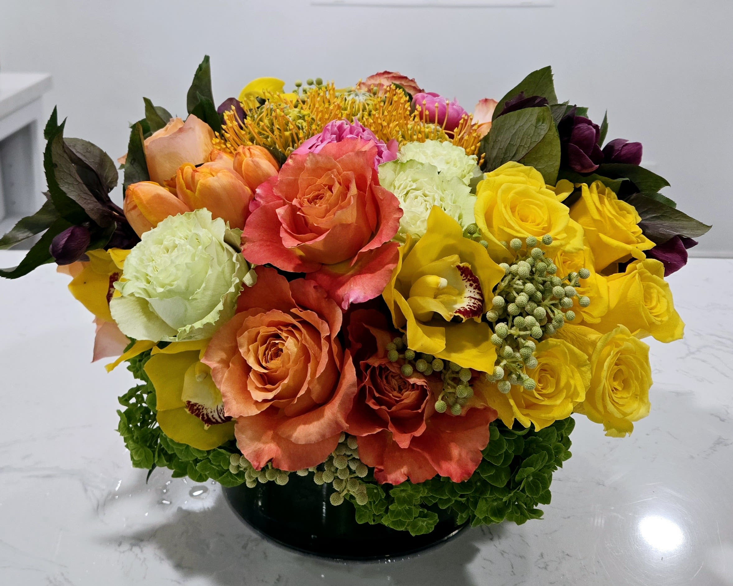 Evening Sun - A vibrant mix of orange, yellow, and peach roses, accented with green hydrangeas and orchids, creates a warm and cheerful display. Perfect for adding a touch of sunshine to any room or occasion.