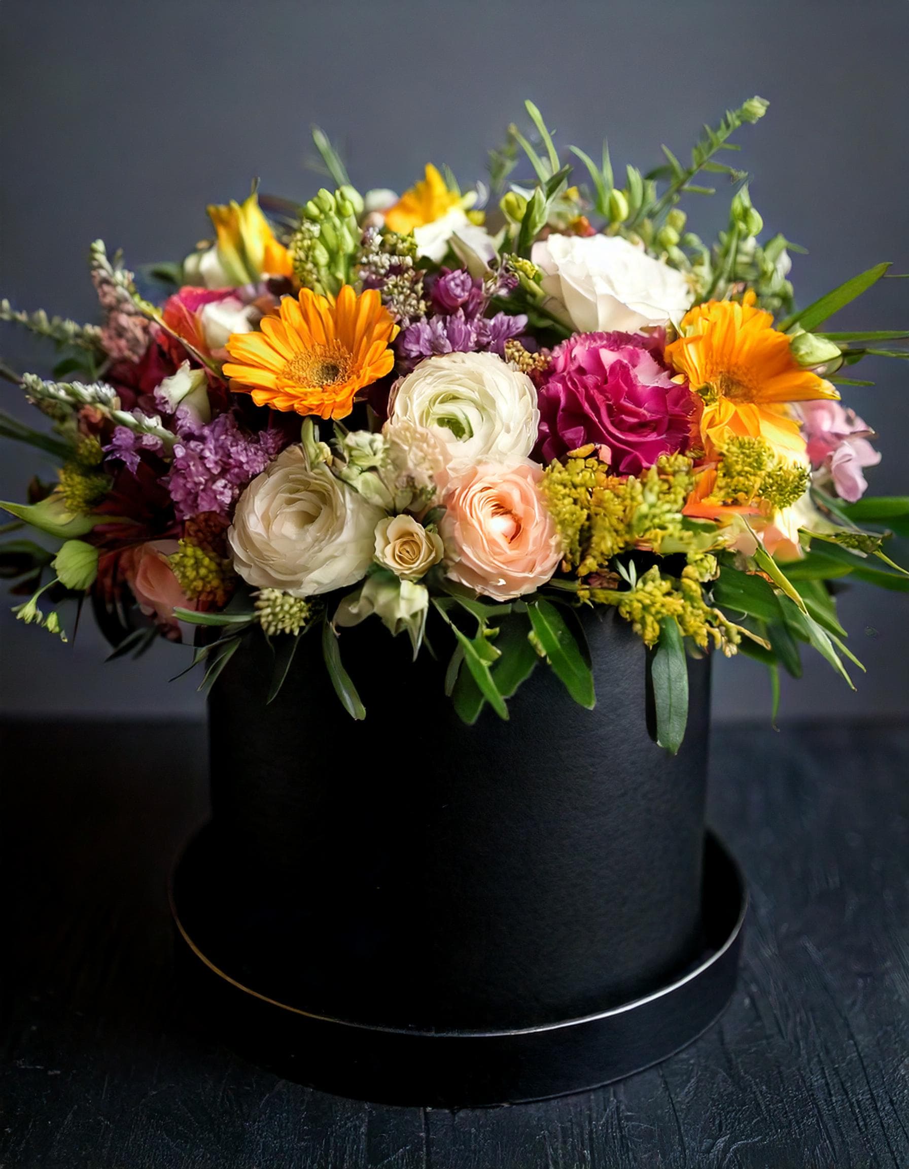 Fresh Vibes - This flower arrangement features of vibrant orange gerberas, delicate white ranunculus, and roses, nestled in a sleek black hatbox. The lush greenery and sprigs of lavender add a touch of elegance, making it a perfect centerpiece for any sophisticated occasion. Elevate your space with this stunning display, ideal for gifting or enhancing your own home decor.