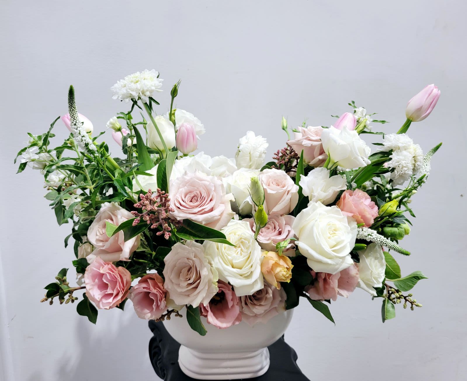 Whispering Blooms - A graceful mix of pale pink and ivory roses, tulips, and lisianthus, beautifully accented by lush greenery. Set in a simple white vase, this arrangement offers a soft and serene elegance, perfect for adding a touch of sophistication to any setting.