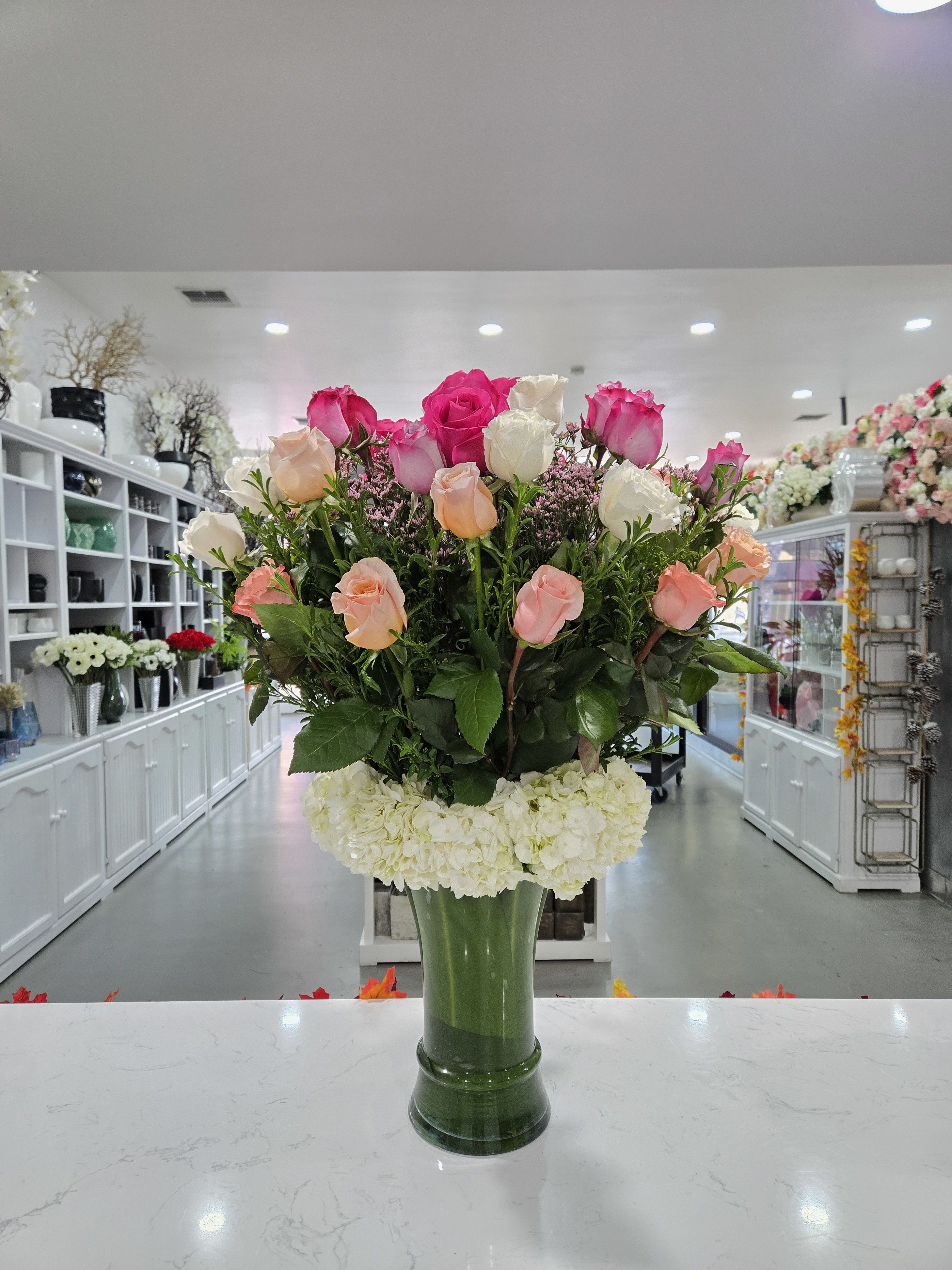 Lizbeth - The Lizbeth arrangement features a mix of pink, white, and peach roses with lush greenery, complemented by white hydrangeas at the base. Presented in a sleek green vase, it offers a classic and elegant look, perfect for any occasion.