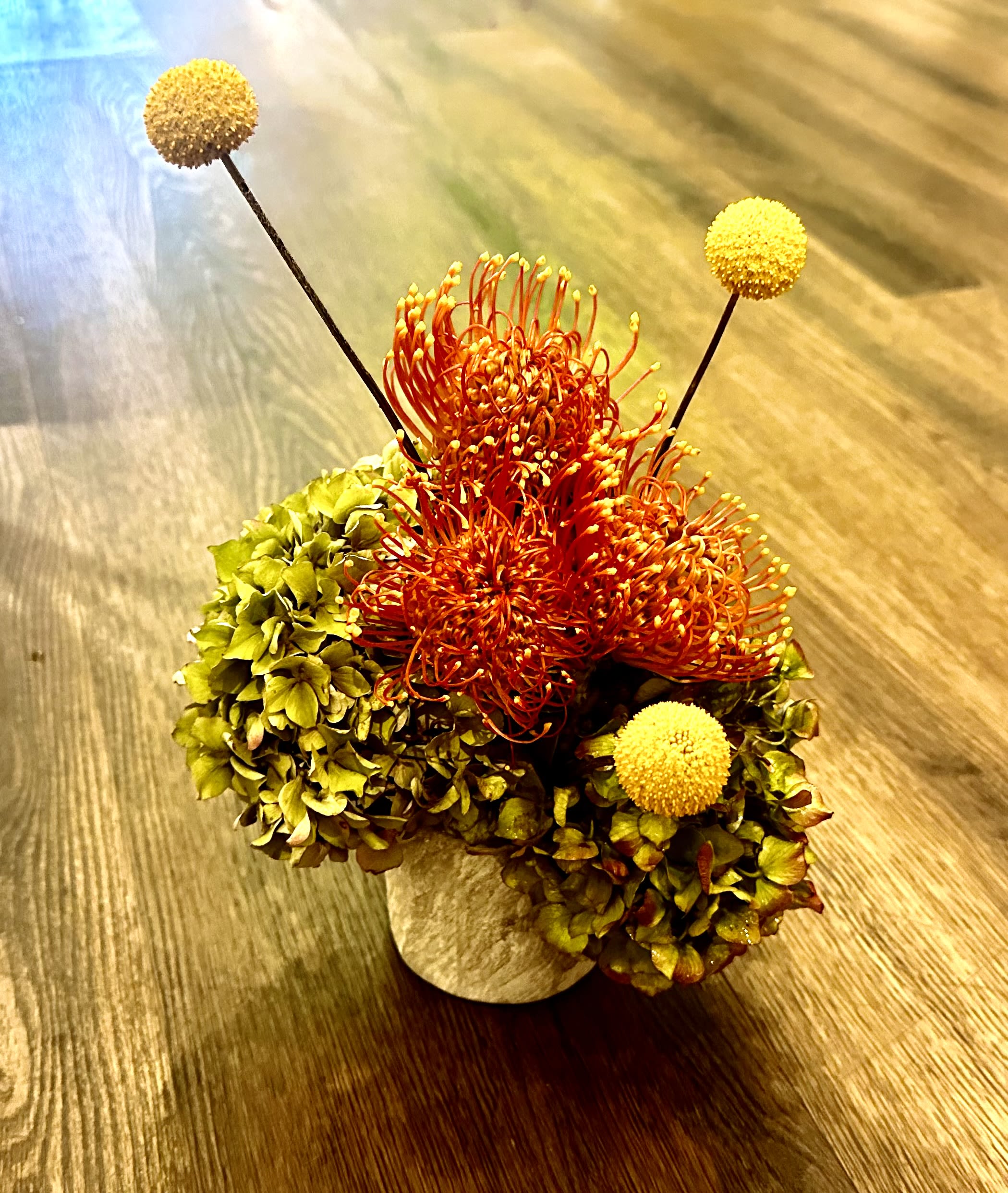 Sunset Serenade - The Sunset Serenade arrangement captures the vibrant hues of a breathtaking sunset. Featuring bold orange pincushion proteas and cheerful yellow billy balls, this piece is harmoniously complemented by lush green hydrangeas. Set in a chic, textured vase, it brings warmth and elegance to any setting. Perfect for adding a touch of nature's beauty to your home or as a thoughtful gift, the Sunset Serenade is available for delivery throughout San Francisco. 