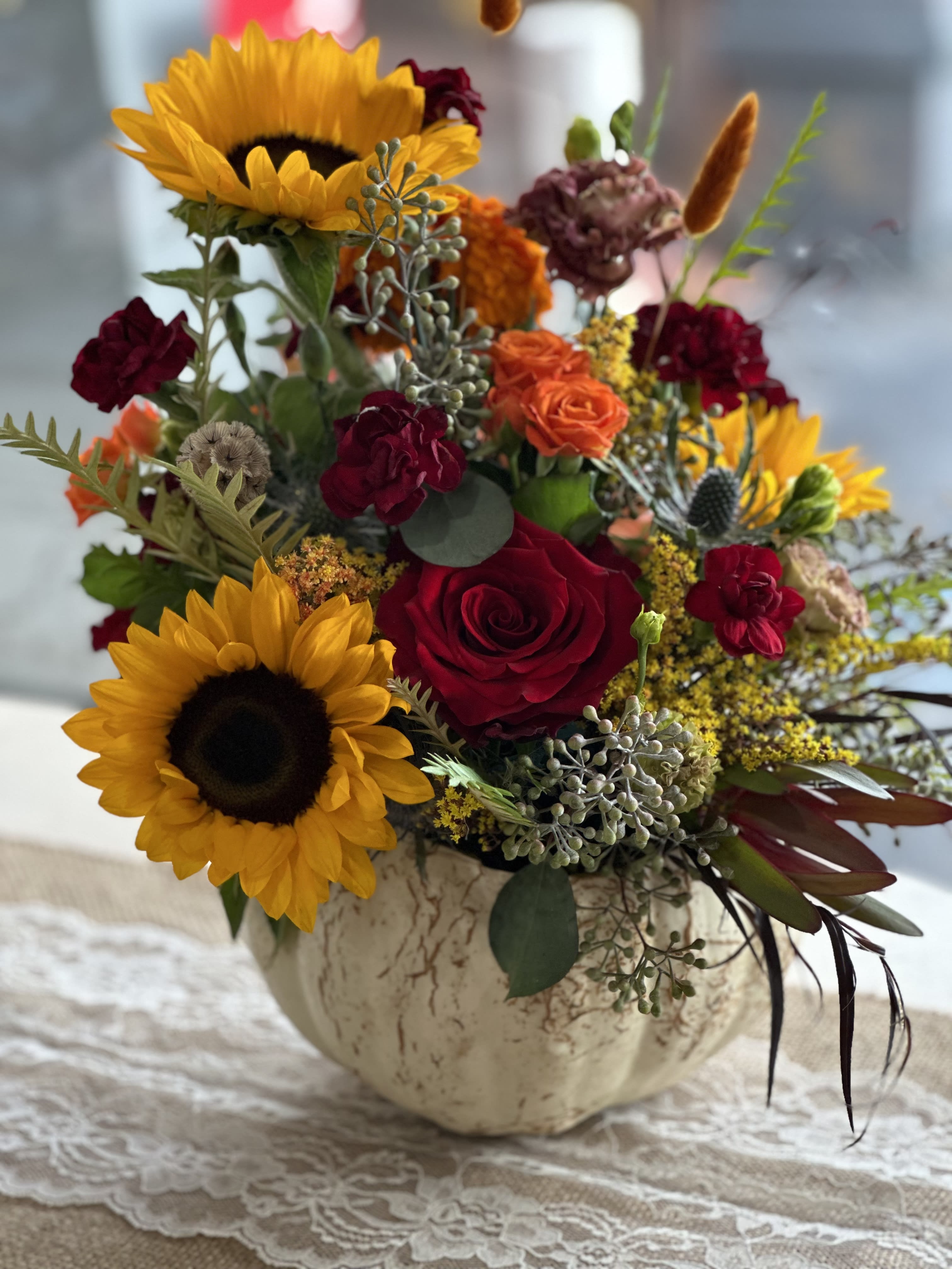 Fall Gourd W/ Sunflowers &amp; Roses  - Elevate your Thanksgiving table this Holiday with our exquisite flower arrangements meticulously curated to capture the rich colors of fall.  Warm tones of burgundy, amber, and deep orange blend seamlessly with vibrant reds and golden yellows that embodies the spirit of the season.   Each arrangement features a beautiful mix of seasonal blooms, complemented by textured foliage and rustic accents.   