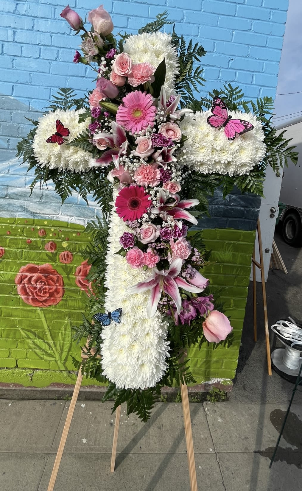 Pink & White Funeral Standing Cross Pink & White Funeral Standing Cross - This funeral standing cross features gerbera daisies, roses, lily’s, carnations, mini carnations, alstroemerias, baby breath, pompon cushion, and a background outline of leather leaf. With three decorative butterflies.