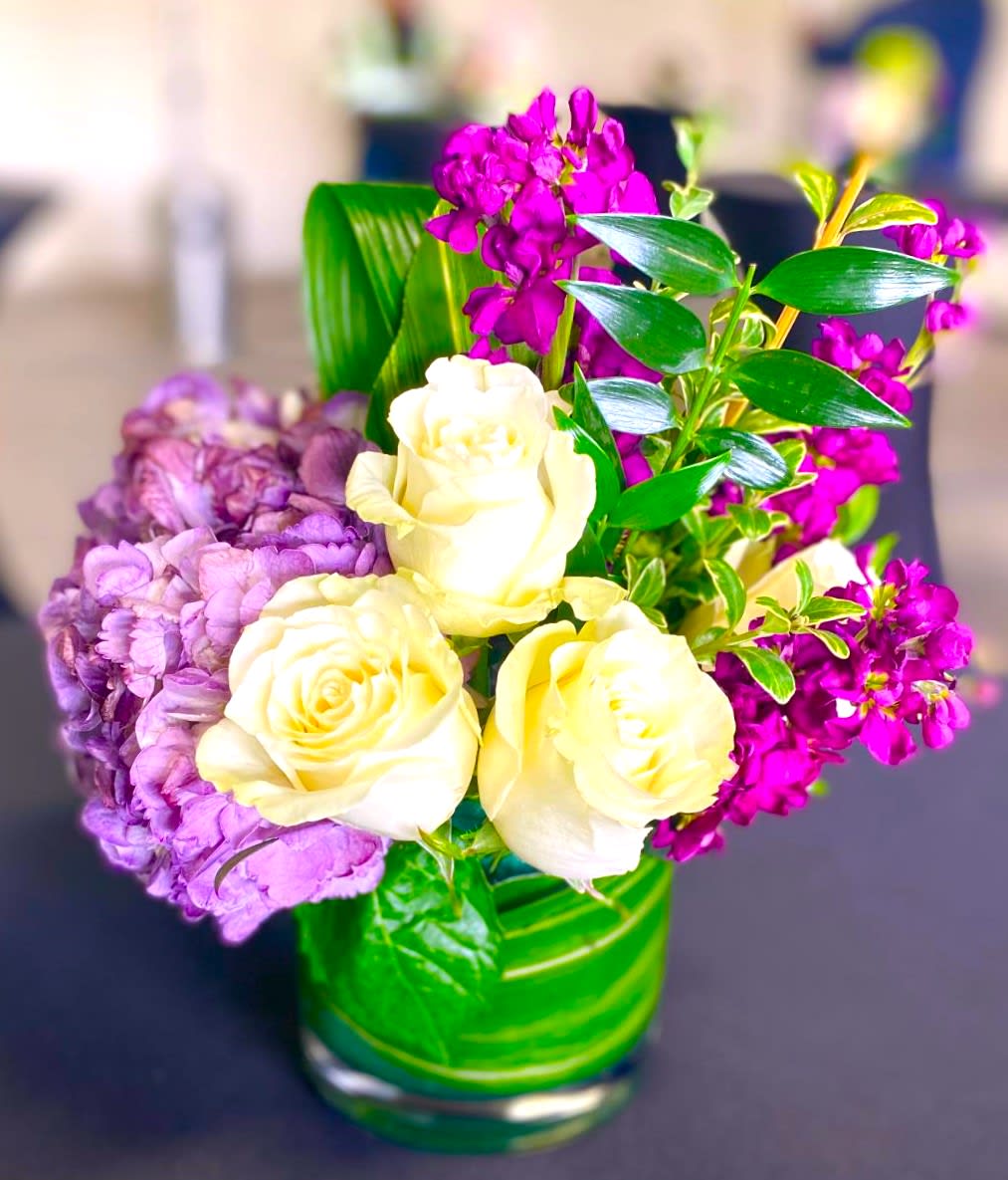 Uptown Love Uptown Love - This simple beauty includes White Roses, Deep Purple Stock, Purple Hydrangea, and Aspidistra leaf ribbons set in a leaf-lined glass cylinder container. This design measures approx. 10"W x 10"H.