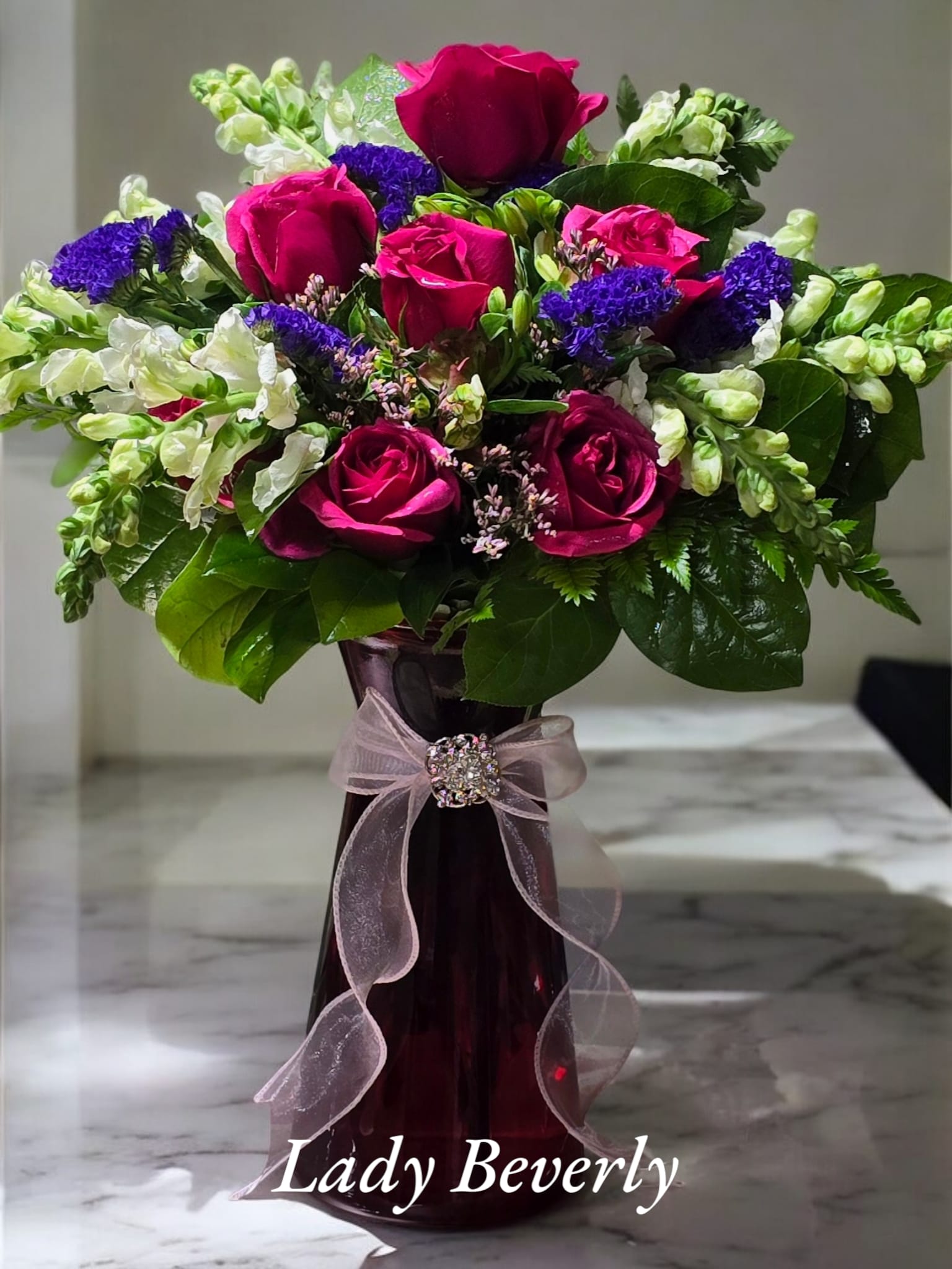 Lady Beverly Lady Beverly - This stunnin arrangement includes Pink roses and white snap dragons. The vase is adorned with a beauriful broche that can be later removed and worn by that special someone.