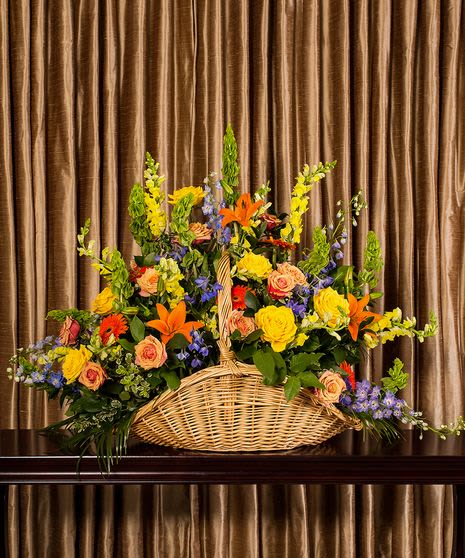 BK # 01    Bright Fireside Basket  - The Bright Fireside Basket is an exquisite floral arrangement, overflowing with life and color, designed to bring a sense of warmth and solace during reflective moments. Its artful composition is both comforting and uplifting, perfect for gracing a fireside where loved ones gather to remember and celebrate a life.  The basket itself is a rustic, woven piece that conjures images of handcrafted tradition, lending an organic touch to the display. It's brimming with an array of flowers: vibrant yellows of roses and snapdragons offer a burst of sunshine, symbolizing friendship and optimism. Oranges, seen in the lilies and gerberas, evoke enthusiasm and passion, a nod to a vivacious spirit. Sprigs of purple and blue delphiniums and hydrangeas add a touch of serenity and depth, providing a beautiful contrast to the warm tones. This floral selection is not just a visual feast but also an aromatic one, with each bloom contributing to a comforting scent that fills the room.