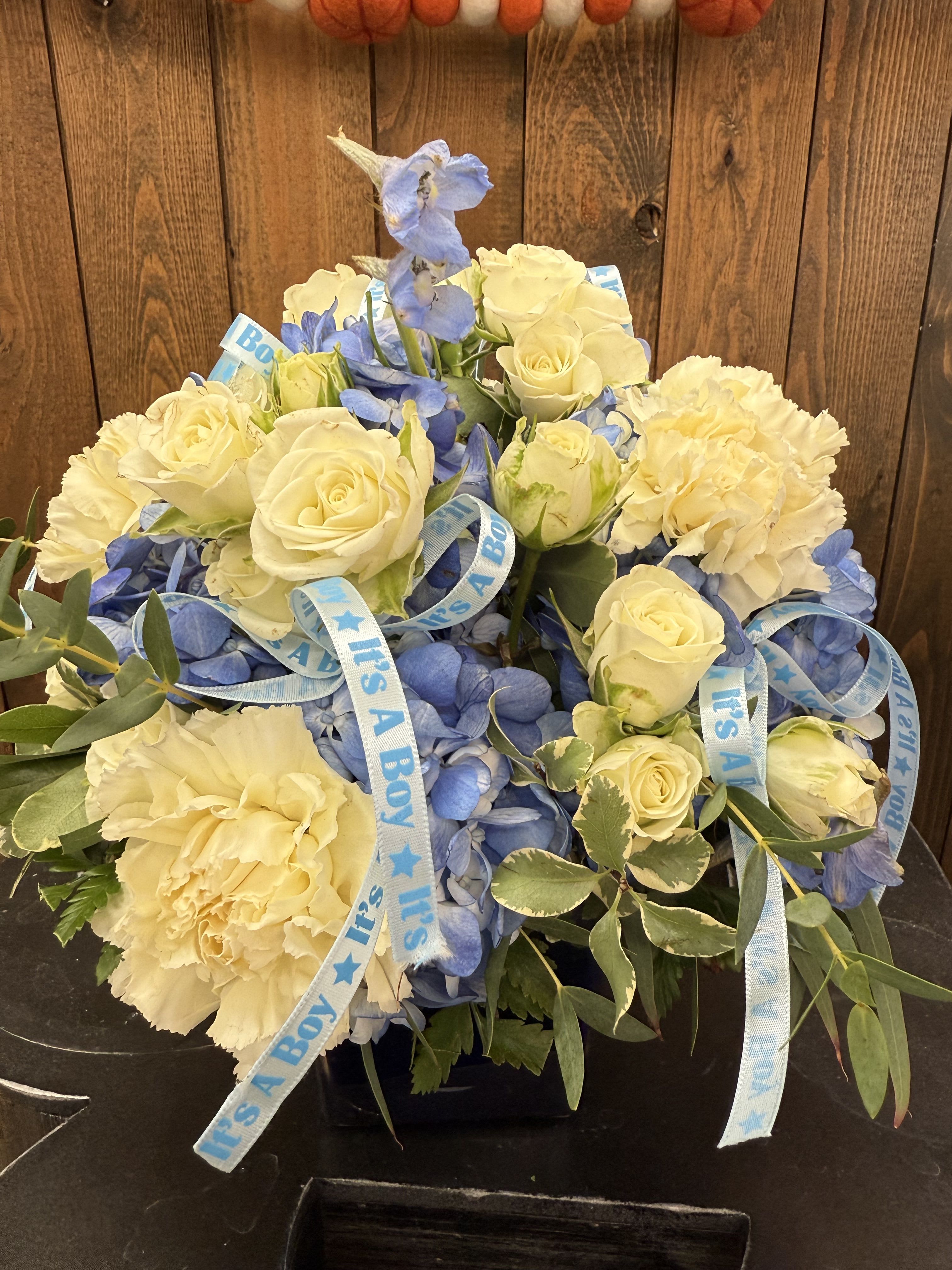 Beautiful Boy - Envision a charming floral arrangement celebrating a baby boy. With beautiful blue hydrangeas, surrounded by white roses and buttery yellow carnations. The arrangement is complemented by lush greenery adorned with a baby boy ribbon making it a perfect centerpiece for welcoming home a new little one.