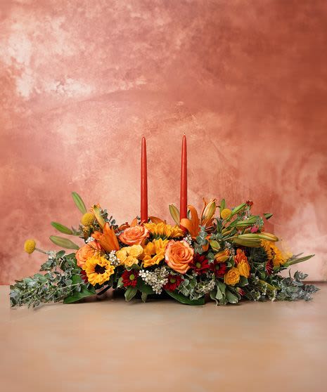 Fall Centerpiece with candle   - The Fall Candle Centerpieces are a radiant and warm arrangement, perfect for any fall gathering. Designed on a classic centerpiece tray, it features a glowing orange candle at its center, casting a cozy ambiance over the table.  Surrounding the candle or candles are vibrant orange lilies, striking burgundy daisy mums, and enchanting Migh and Magic roses, creating a rich, autumnal color palette. Cheerful sunflowers brighten the centerpiece, while elegant Fuji bronze mums add depth and texture. Delicate wax flowers provide softness and contrast, enhancing the arrangement’s intricate beauty.