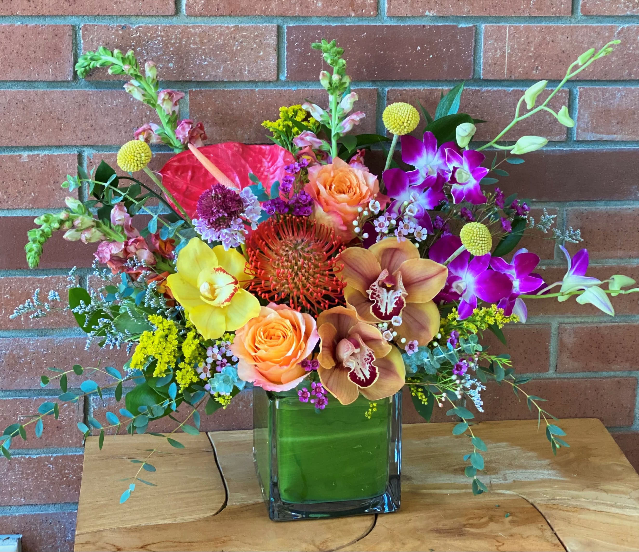 Floridian Sunset  - Arrangement full of vibrant blooms: Orchids, dendrobium purple orchids, orange garden roses, snapdragons, pin cushion protea, anthurium, billy balls, scabiosa, wax flowers, premium greens and fillers arranged in a 5x5x5 square glass vase wrapped with green TI leaves. Standart size shown in the picture 