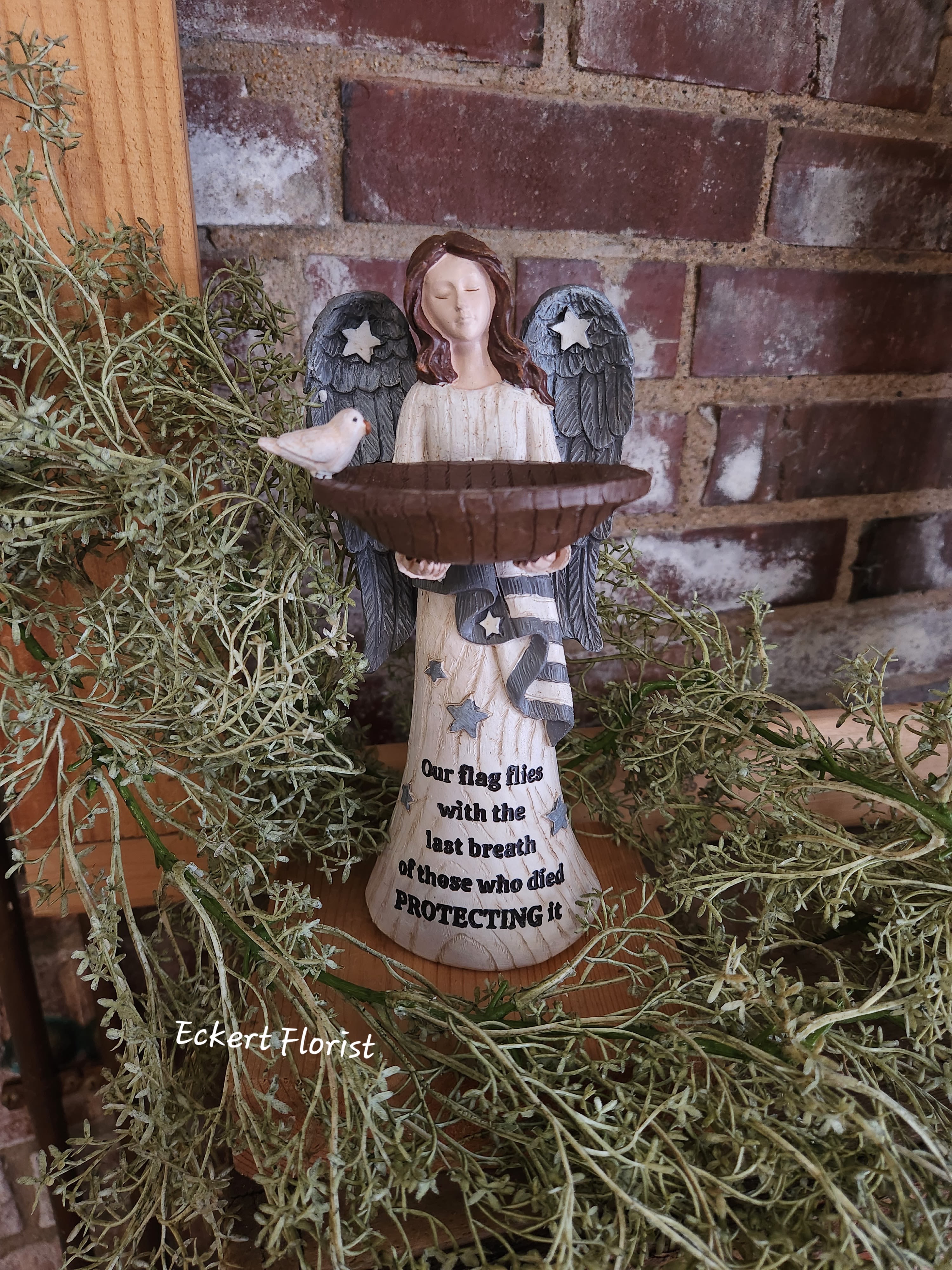 Eckert Florist's "Our Flag Flies..." Angel *Our Local Delivery Only Eckert Florist's "Our Flag Flies..." Angel *Our Local Delivery Only - Resin angel measures approx. 8.5" H x 3.75" wing span. Sentiment: "Our flag flies with the last breath of those who died Protecting it." Can be added to a green plant or fresh arrangement. See Deluxe or Premium upgrades