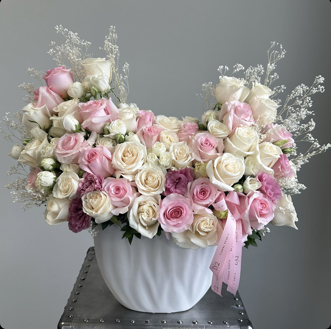 Pink Monica - This arrangement features a delicate mix of soft pink and white roses, creating a romantic and serene display. The roses are accompanied by baby's breath, which adds a whimsical, airy touch to the overall design. The arrangement is housed in a smooth, white ceramic vase, which complements the pastel tones of the flowers. A pink ribbon is delicately draped over the front, adding a charming detail. The arrangement exudes elegance and is perfect for celebrating special occasions like baby showers, birthdays, or anniversaries.
