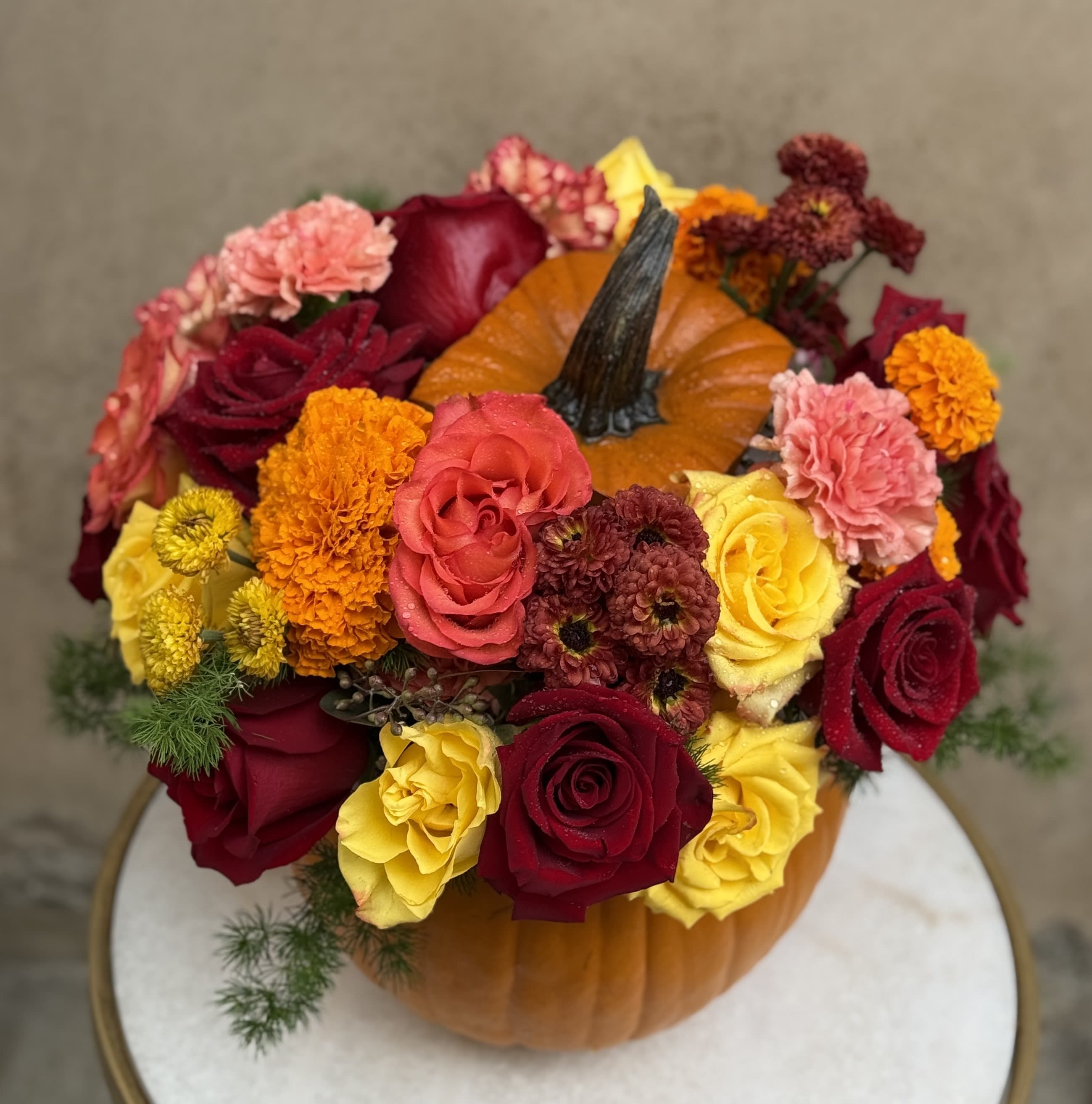 Autumn Glow Pumpkin Arrangement - Embrace the warmth of autumn with our Harvest Pumpkin Blossom Bouquet, a vibrant mix of seasonal blooms arranged in a festive pumpkin vase. This delightful arrangement may consist of but not subject to deep red roses, cheerful yellow roses, bright marigolds, and soft pink carnations, all nestled around a real pumpkin for an extra touch of fall charm. Perfect as a centerpiece for your Thanksgiving table or as a gift to bring autumnal joy to someone special. Celebrate the season with this cozy and colorful pumpkin arrangement that captures the spirit of fall! 