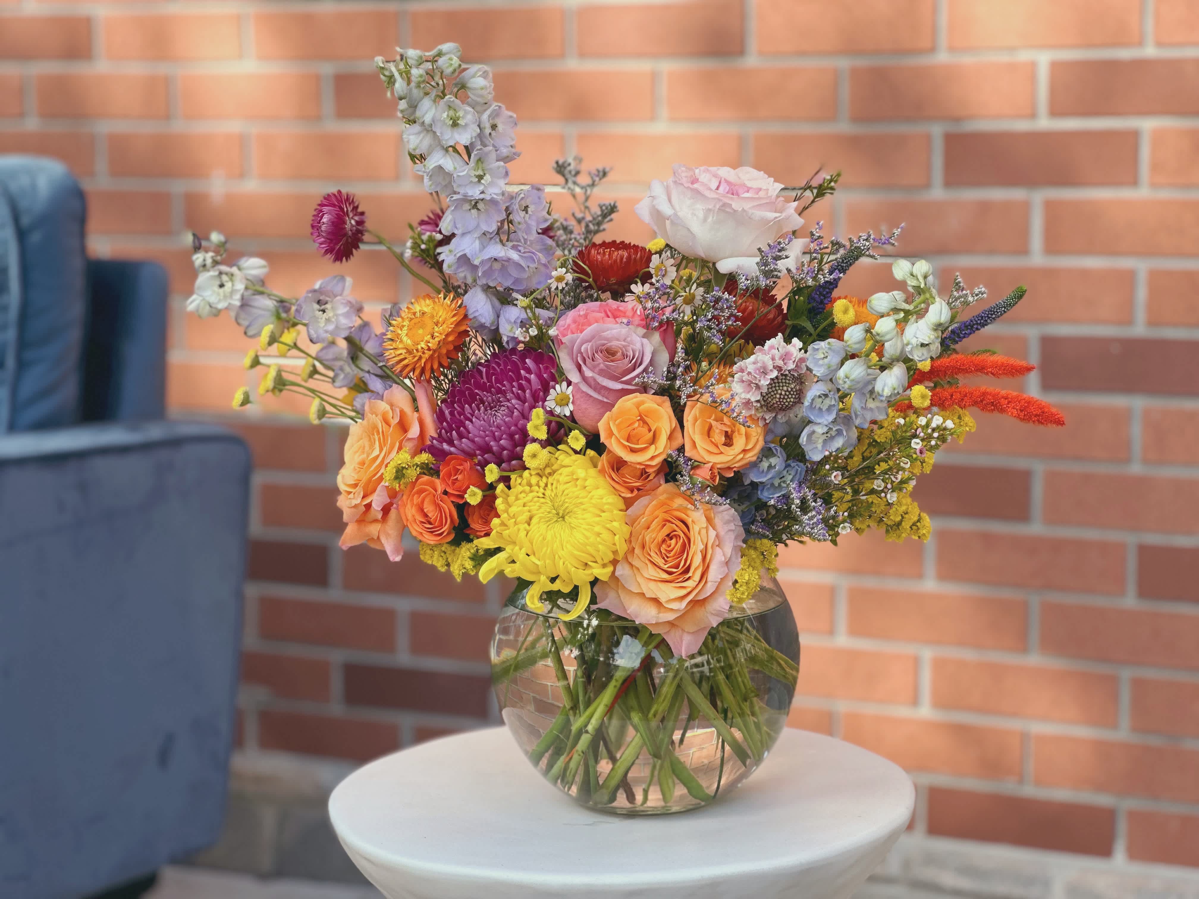 Bubble Of Wildflowers   -  The bouquet in a bubble blown is a unique and whimsical arrangement filled with a vibrant mix of wild flowers. The delicate blue delphinium stands tall and proud, complemented by soft garden roses and delicate scabiosa. Veronica adds a touch of elegance with its tall spires, while solidago and lumonium provide a pop of sunny yellow.  The strawflowers add a fun and playful element to the bouquet, while the delicate ranunculus bring a touch of romance. Each flower seems to dance and sway within the bubble blown, creating a magical and enchanting display.  Overall, this bouquet is a stunning mix of wild flowers that showcases the beauty and diversity of nature. It is a true work of art that will bring joy and happiness to anyone who beholds it.