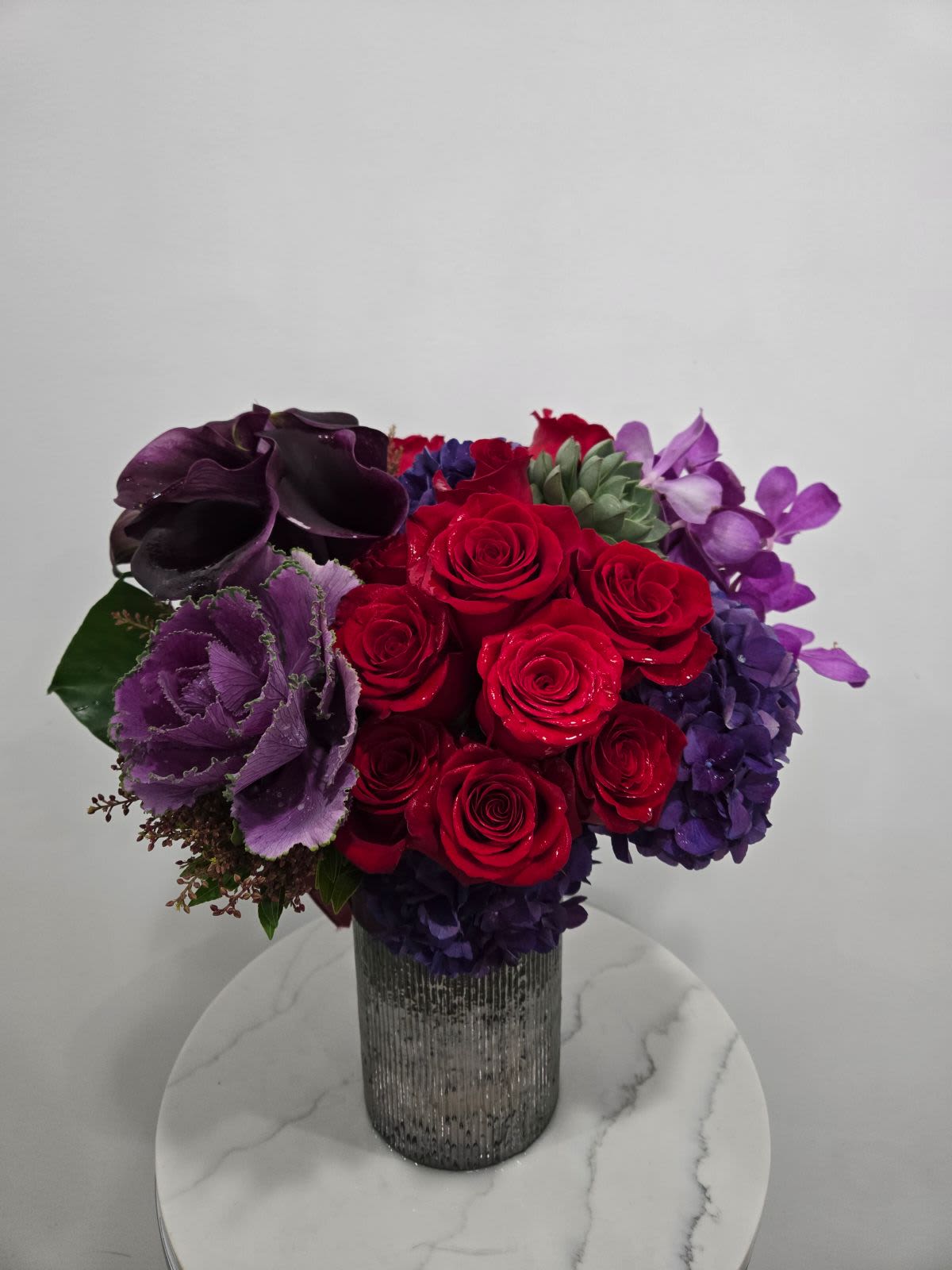 Regal Bloom - A luxurious and bold floral arrangement featuring vibrant red roses, deep purple calla lilies, and delicate succulents, complemented by rich purple hydrangeas and ornamental kale, all set in a textured silver vase. Perfect for making a statement or adding elegance to any space.