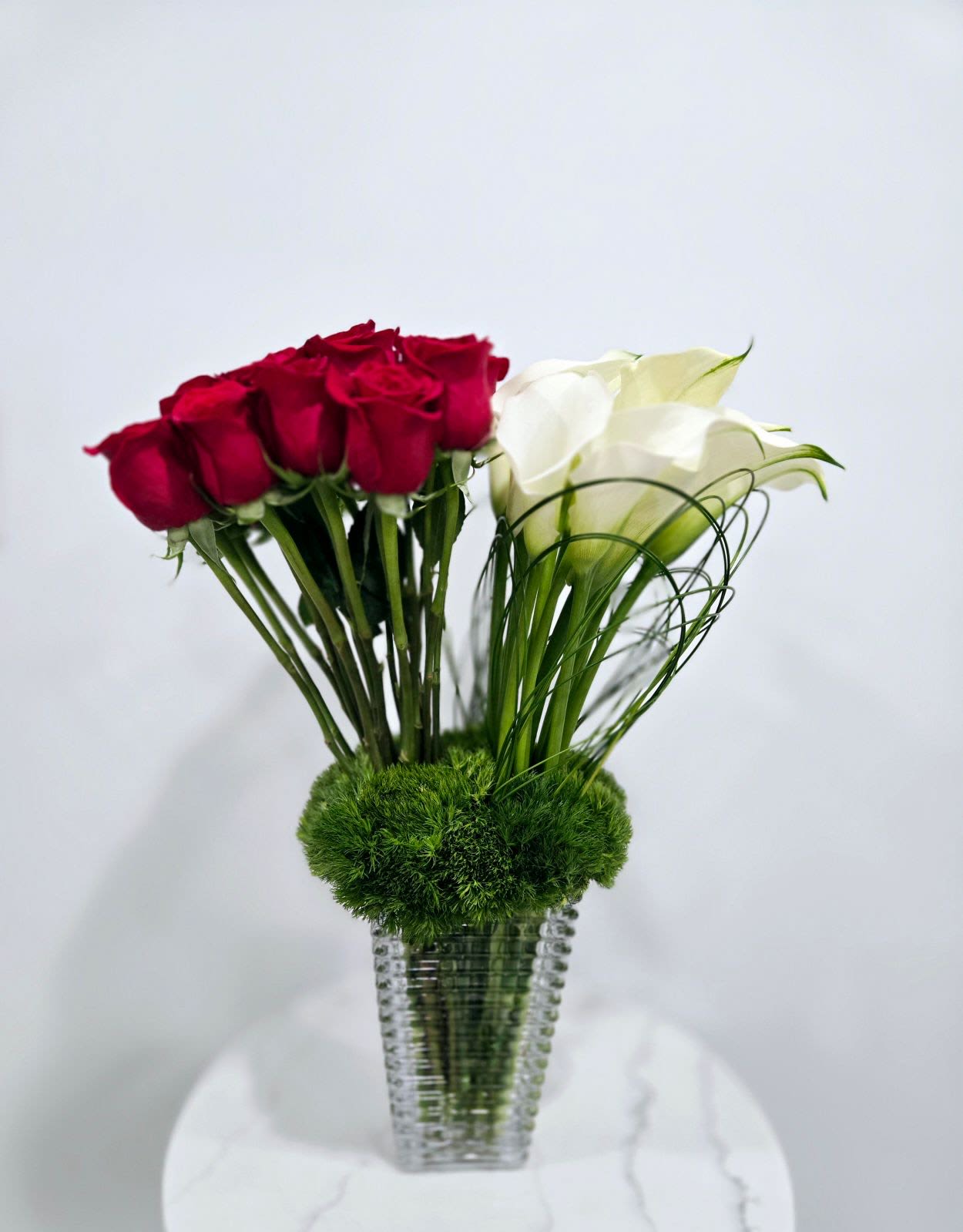 Urban Romance -  A sleek fusion of red roses and white calla lilies anchored in lush green moss, presented in a contemporary textured glass vase. This arrangement captures modern elegance with a hint of bold flair.