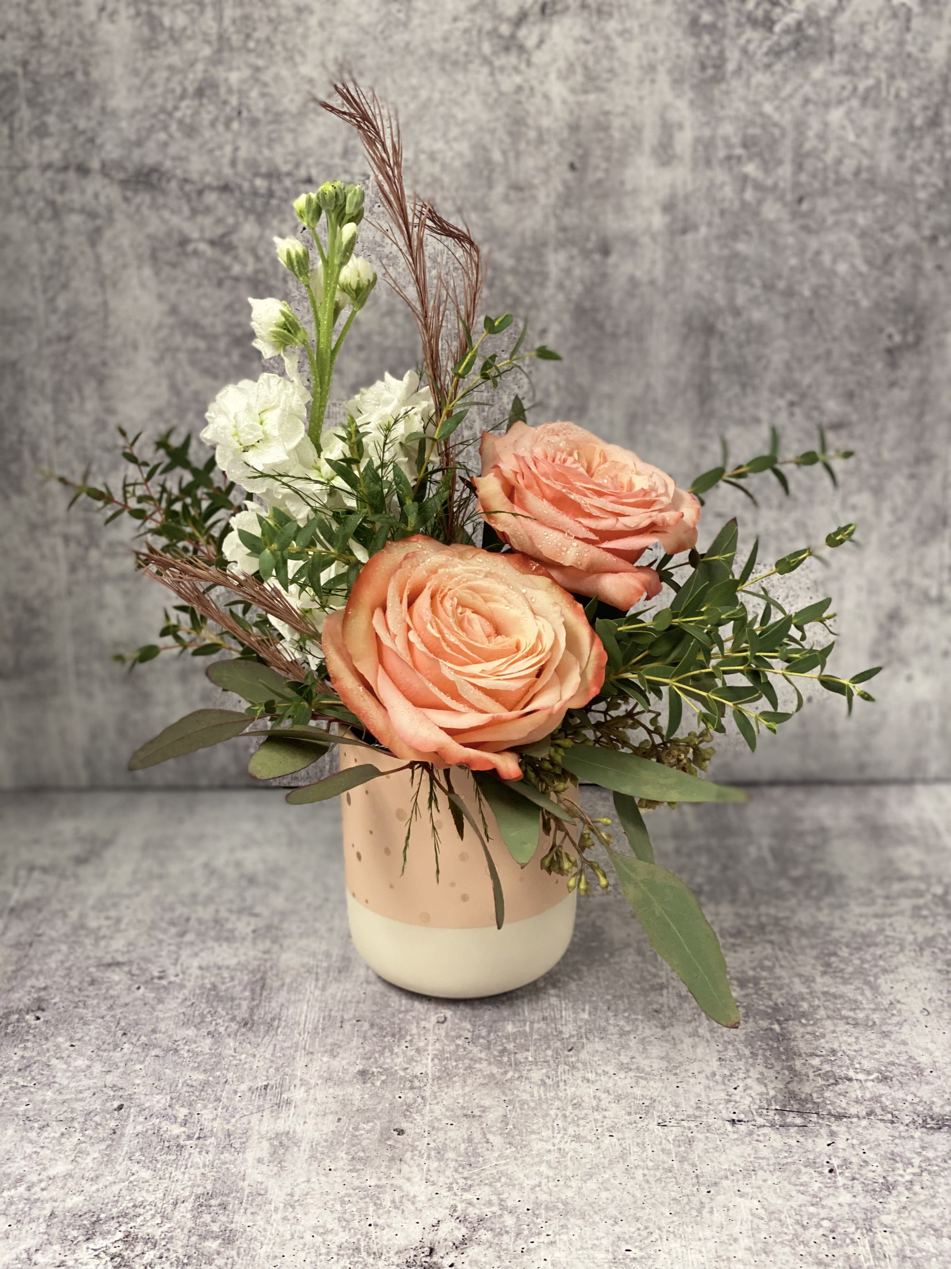 Sweet Nola - Kahala roses with Eucalyptus greens, sensationally scented white Stock and preserved Hazelnut Asparagus in a white and apricot/light pink container.