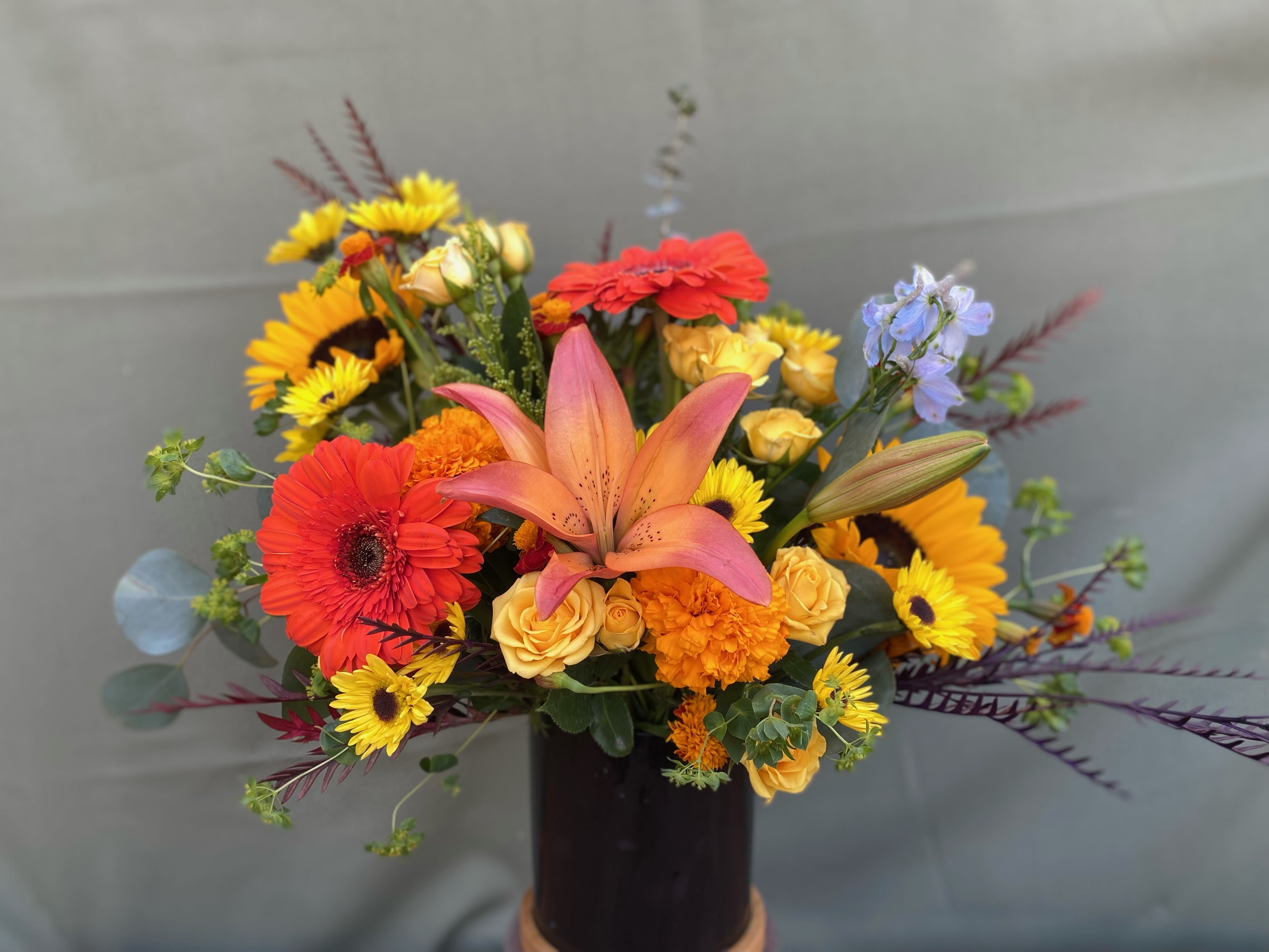 Cutie Cutie - This arrangement includes Gerbera daisies, tiger lilies, sunflowers, mini spray yellow roses, blue delphinium, all hand arrange in black ceramic vase.