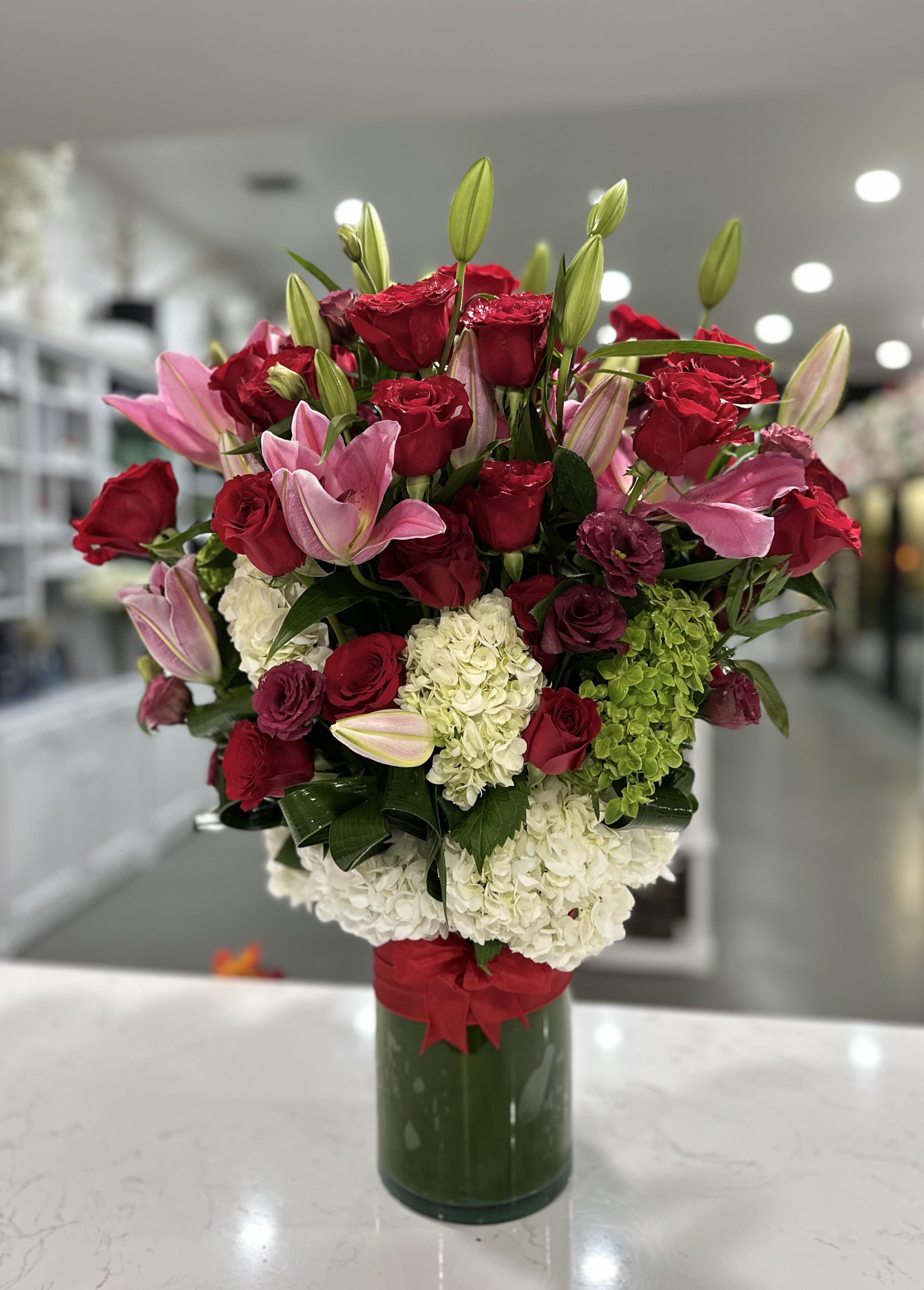 Touch of Love - This elegant floral arrangement features a lush display of red roses, pink lilies, white hydrangeas, and touches of green accents, beautifully arranged in a cylindrical green vase wrapped with a red ribbon. The mix of colors and textures creates a vibrant and sophisticated look, perfect for a romantic or celebratory setting.