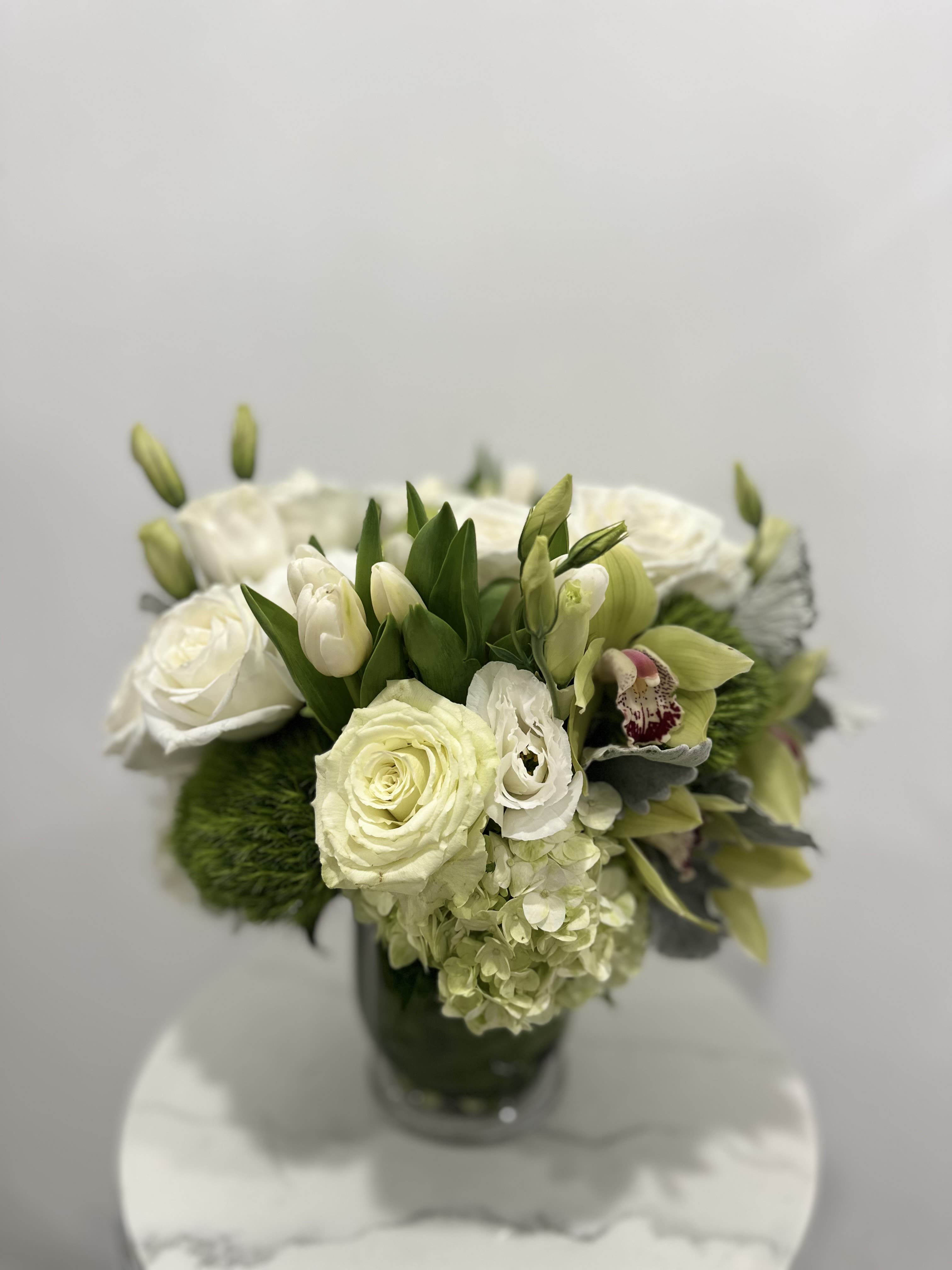 Verdant Elegance - This exquisitely arranged bouquet radiates serene beauty and sophisticated charm. It features a stunning collection of white roses and tulips accented with verdant hydrangea blossoms and the textured intrigue of green chrysanthemums. Nestled amidst lush greenery, these blooms are elegantly displayed in a simple, clear glass vase, creating a display of purity and freshness that brings a touch of refined tranquility to any setting. Perfect for special occasions or as a gesture of heartfelt sentiment.