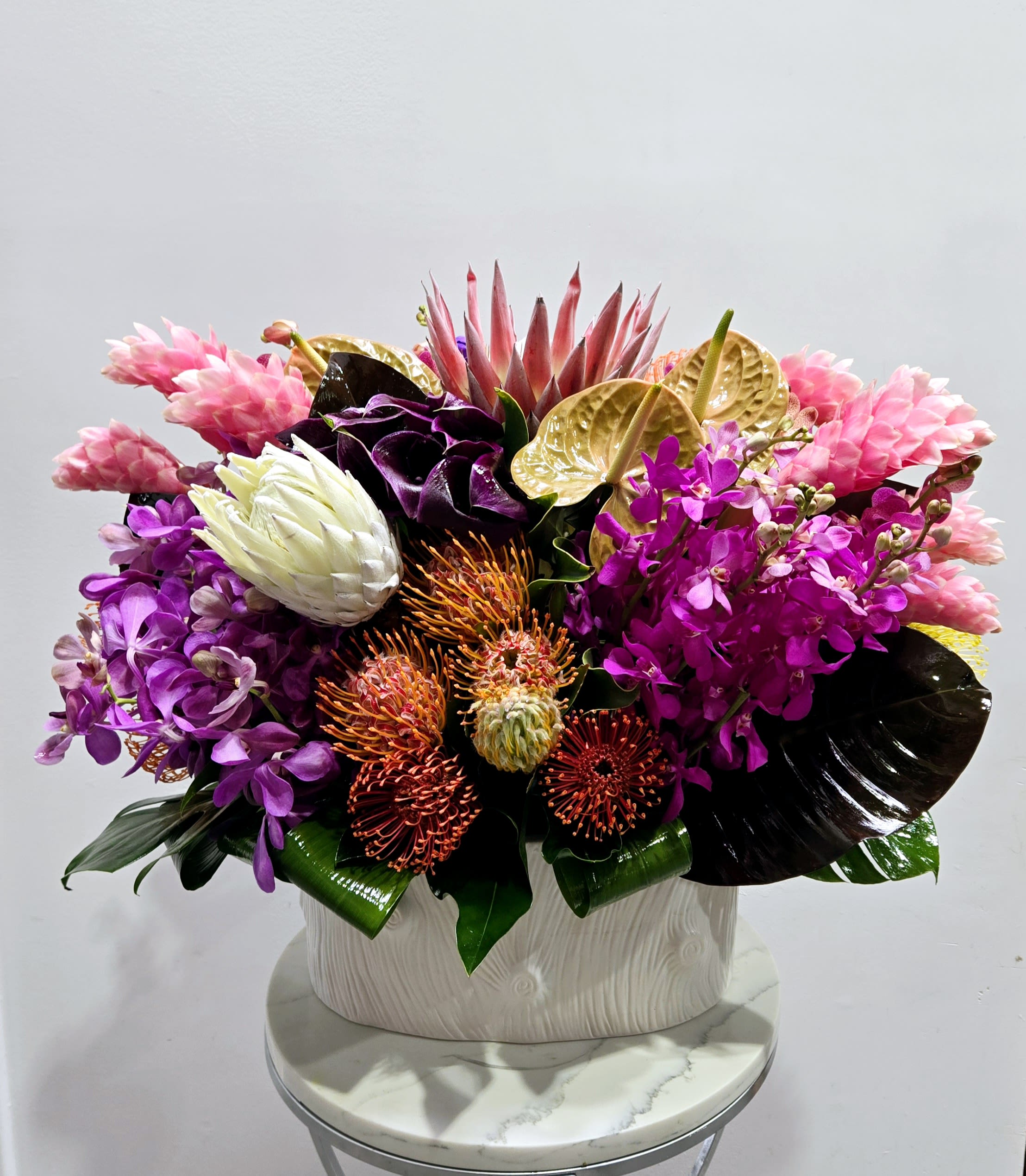 Vivid Tropics - A bold mix of exotic blooms, including pink ginger, purple orchids, and orange pincushion proteas, highlighted by dramatic tropical leaves. This vibrant, tropical arrangement adds a burst of color and energy to any space, perfect for making a striking statement.