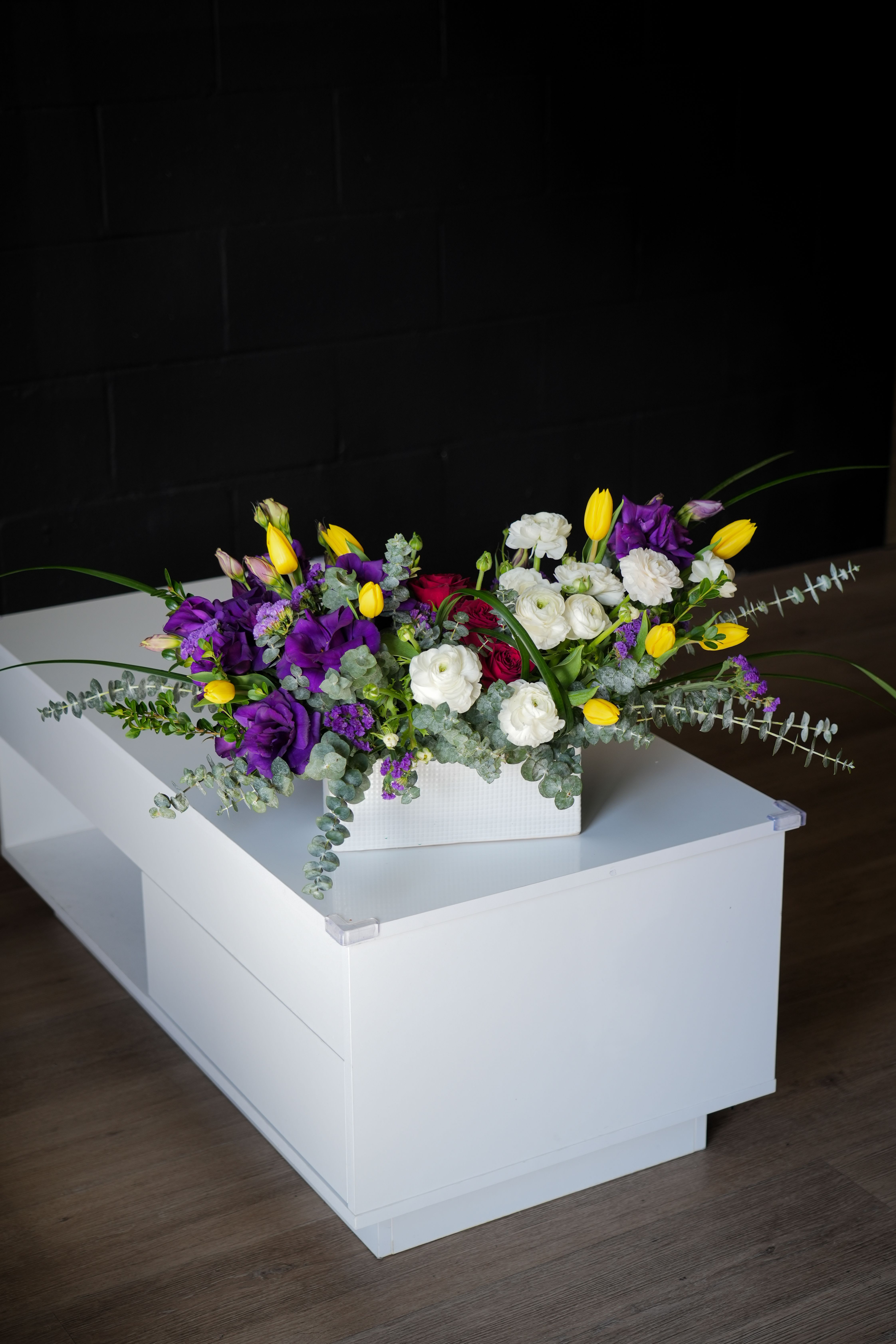 Spring Symphony Arrangement - Brighten any space with the vibrant hues of the Spring Symphony Arrangement! This eye-catching piece combines rich purple blooms, delicate white ranunculus, cheerful yellow tulips, and deep red roses, all complemented by soft eucalyptus leaves for a touch of greenery. Arranged in a sleek white rectangular box, this composition brings an elegant yet playful energy, making it perfect for celebrations, gifts, or as a stylish decor piece. 