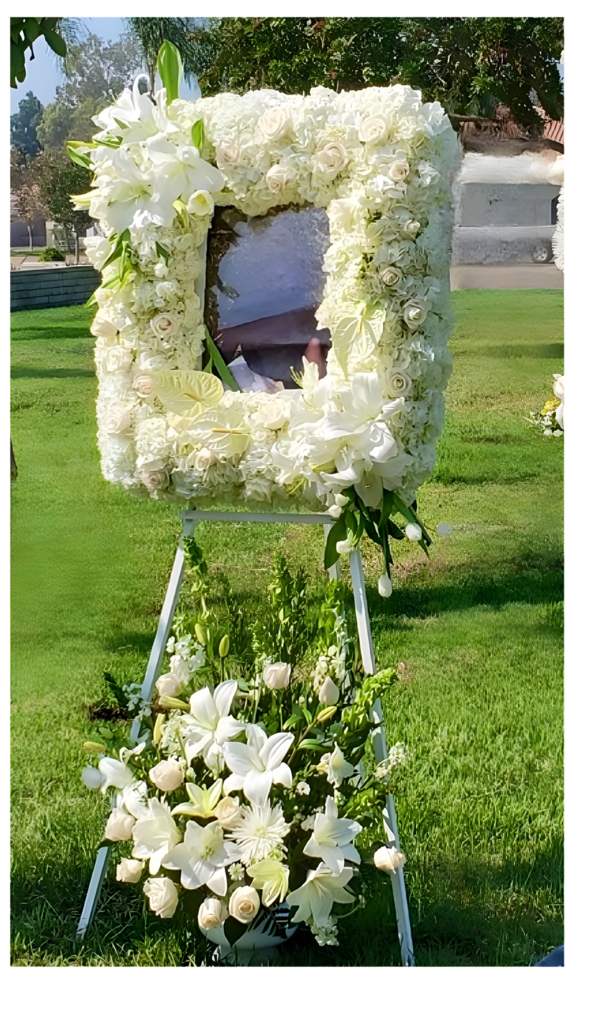Heavens Portrait  - This exquisite, handcrafted floral tribute is a touching way to honor the memory of a loved one.  Designed in a boutique style, this unique piece features a  photo frame surrounded by a cascade of pristine white blooms, symbolizing purity, innocence, and sympathy.  Each tribute is made to order with the freshest lilies, roses, carnations, anthurium, and orchids, and is securely mounted on an easel for elegant display.  Perfect for a funeral, memorial service, or graveside remembrance, this bespoke creation offers a personal and heartfelt touch. It serves as the perfect complement to an urn or casket spray.  Features:  Handcrafted with fresh flowers Boutique style design White flowers symbolize purity and sympathy Includes a photo frame to display a cherished picture Mounted on an easel for easy display Made to order Perfect for funerals, memorial services, or graveside remembrances Complements urn or casket sprays Matching basket can be added to the base of the easel (sold separately)