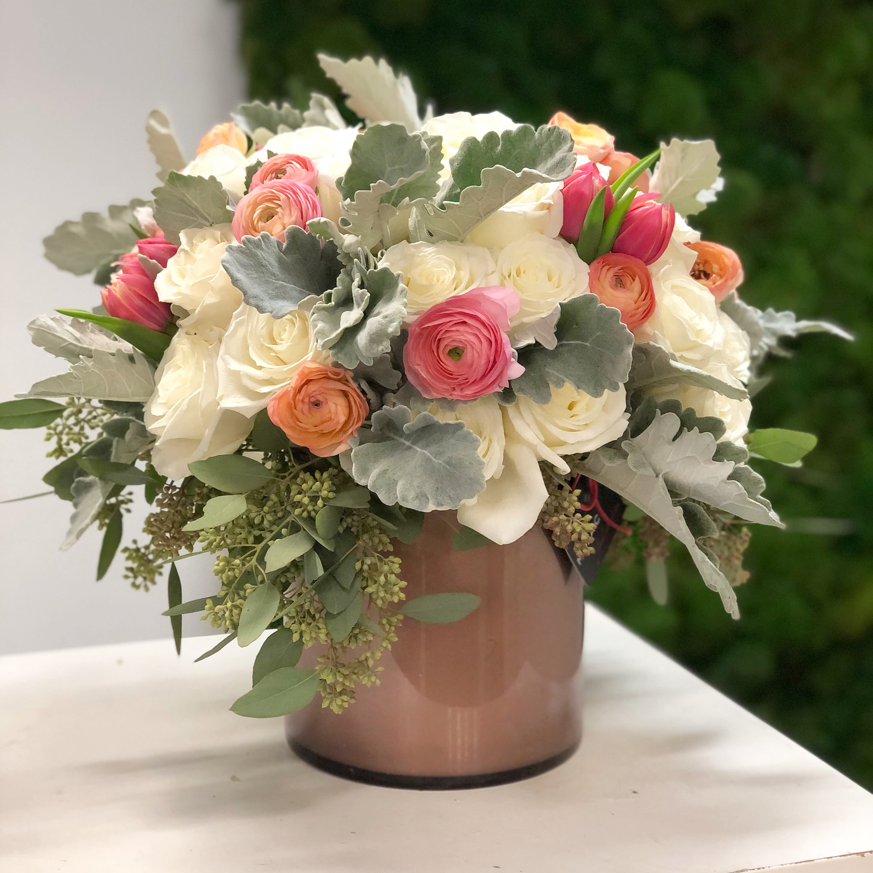 Muted Muted - A timeless blend of soft hues and textures, this arrangement features lush white roses, delicate peach and pink ranunculus, and vibrant tulips nestled among silvery dusty miller and cascading eucalyptus. Arranged in a sleek, blush-toned vase, this design is perfect for adding a touch of refined style to any space or occasion. Ideal for celebrations, gifting, or simply elevating your home décor. Dimensions: Approximately 12" H x 10" W Care: Keep in a cool area, away from direct sunlight and AC. Replenish water every 2-3 days. Available for delivery or in-store pickup.