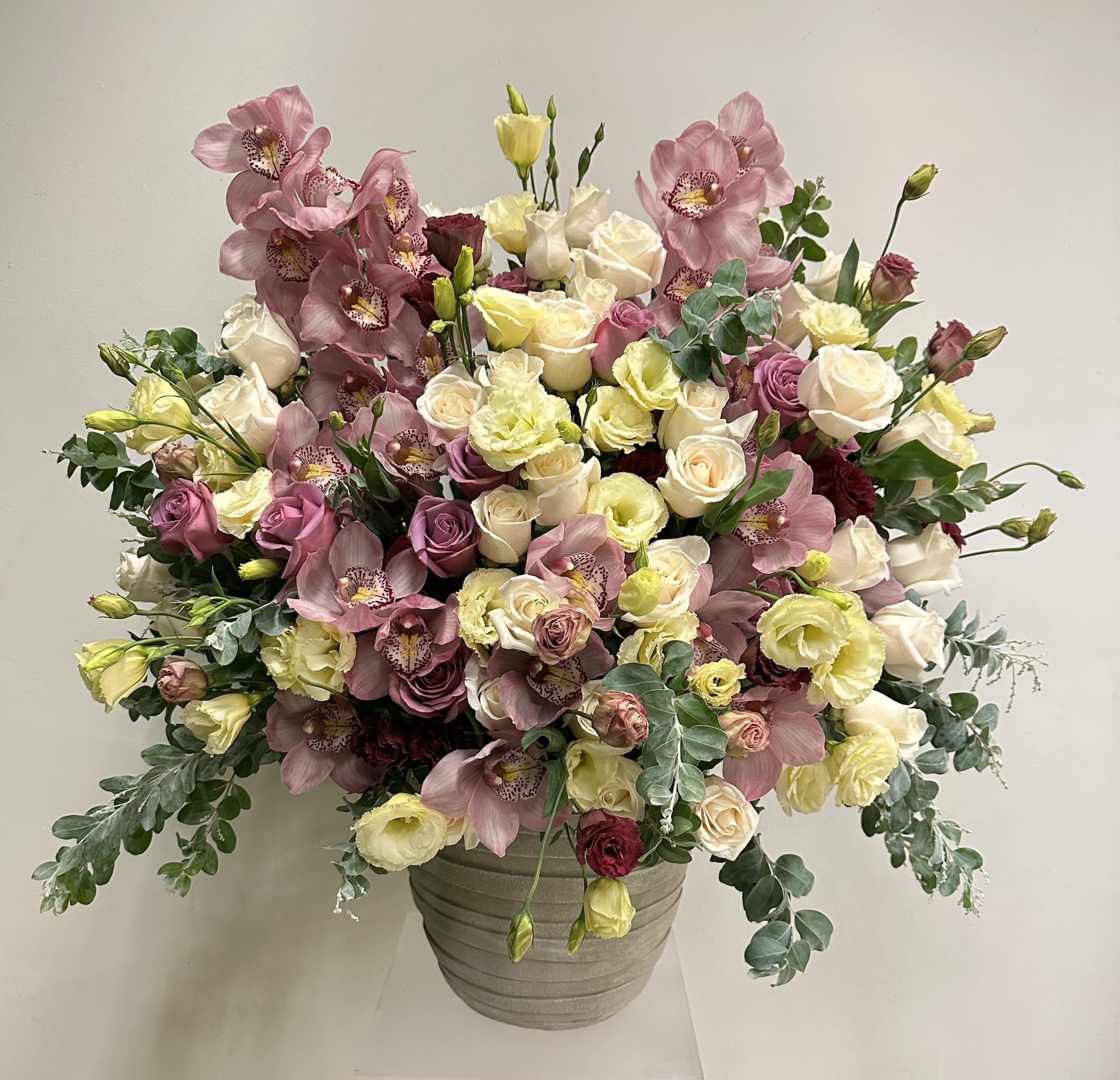 Floral Explosion Floral Explosion - A luxurious blend of soft cream, dusty lavender, and blush hues, featuring elegant roses, delicate lisianthus, and striking cymbidium orchids. Accented with lush eucalyptus foliage, this arrangement radiates sophistication and timeless beauty. Perfect for making a bold statement in lobbies, dining areas, or special celebrations. Dimensions: Approximately 3 feet tall and 2.5 feet wide, designed in a textured ceramic vase.