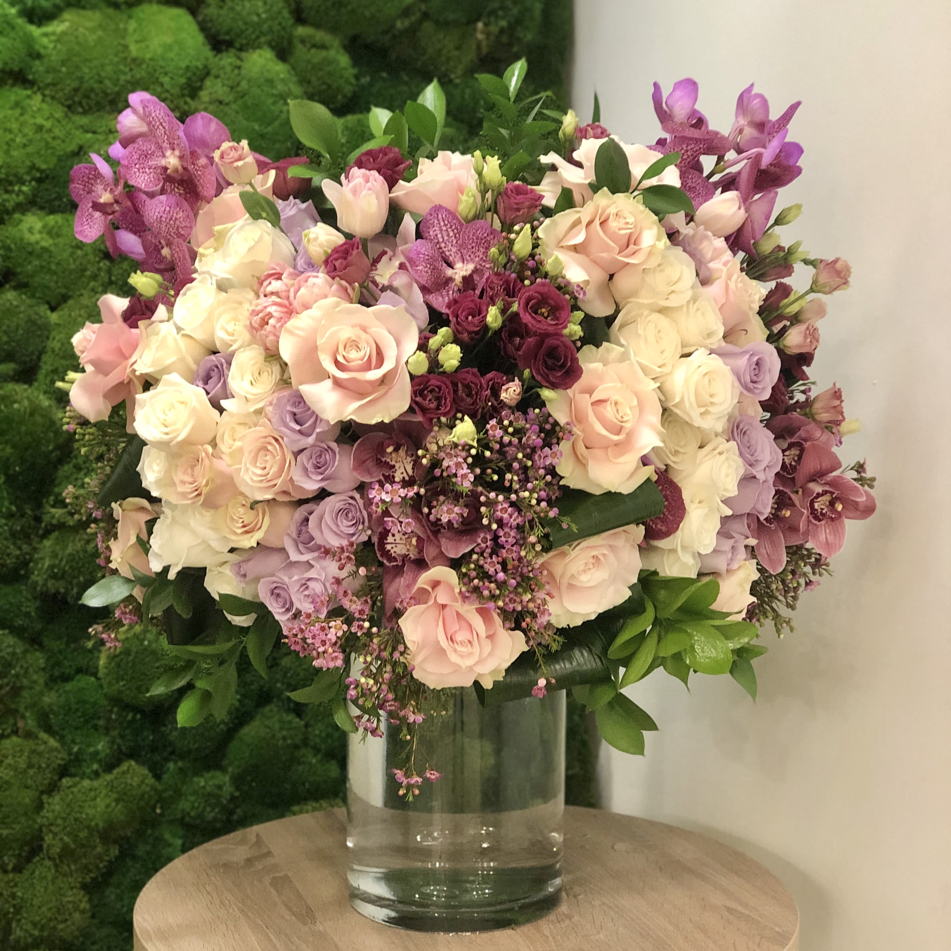 French Garden French Garden - This elegant floral arrangement showcases a delicate blend of pastel roses in soft pink, ivory, and lavender hues, complemented by deep plum lisianthus, vanda orchids, wax flowers, and cymbidium orchids. Accentuated with fresh greenery, all blooms are thoughtfully arranged in a timeless clear glass vase, making it a perfect statement piece for weddings, anniversaries, or any sophisticated occasion. A design that exudes romance and refined beauty. Overall height is 30"